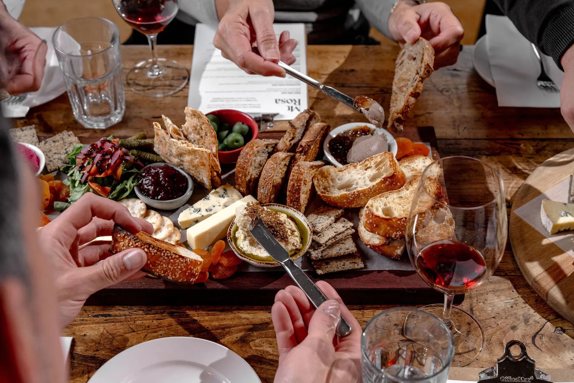 Group enjoying cheese board with meats, bread, olives, and wine during winery tasting experience on wooden table.