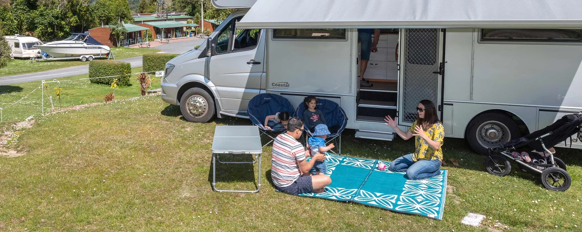 Holiday park with family relaxing beside motorhome, outdoor mat, chairs and pram on grass camping site.
