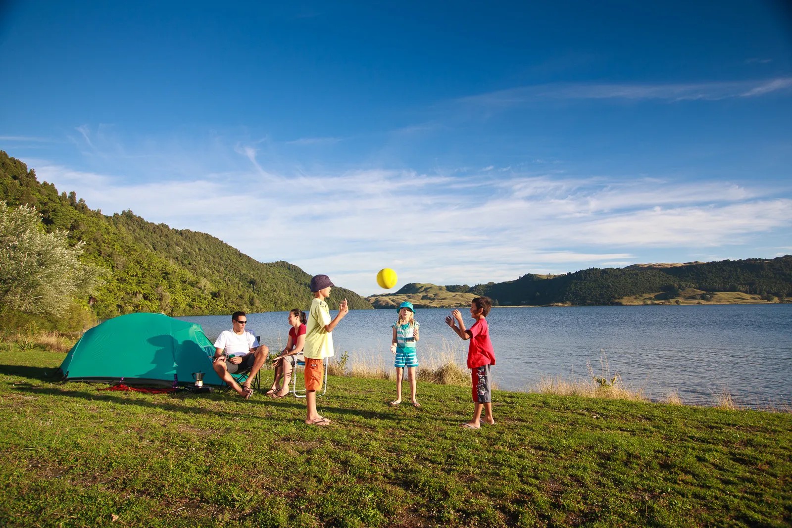 Lake Okareka Campsite with family camping by green tent, children playing and scenic lake and hills backdrop.