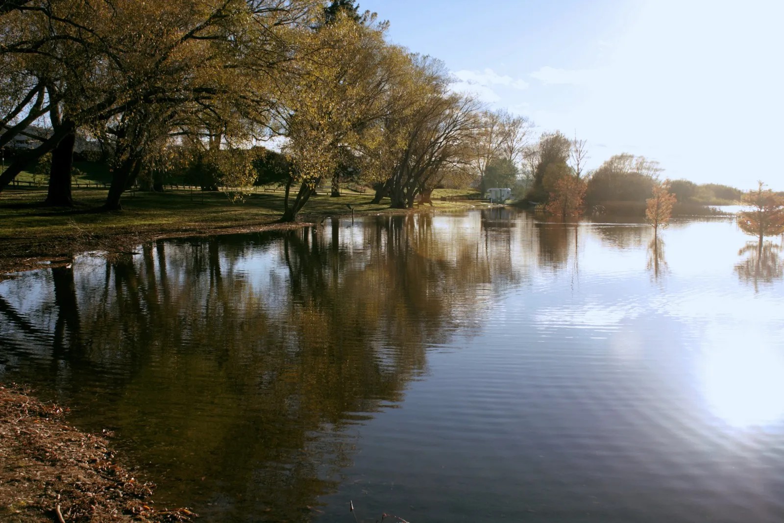 View of Lake Rerewhakaaitu with orange-leaved trees reflected in the water and a motorhome on the land on the far side of the lake.
