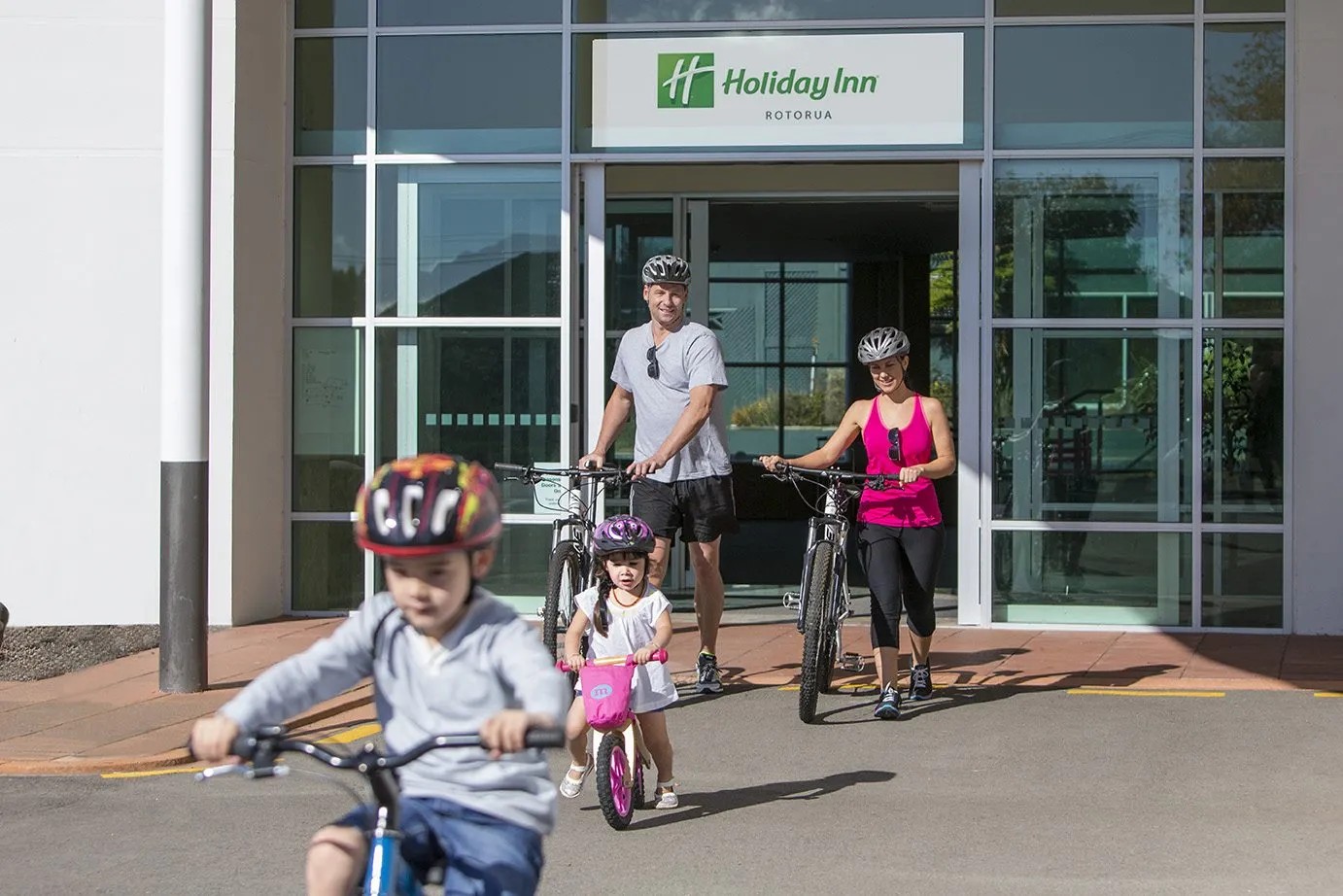 Family with bicycles outside Holiday Inn Rotorua entrance, parents and children wearing helmets ready for cycling.