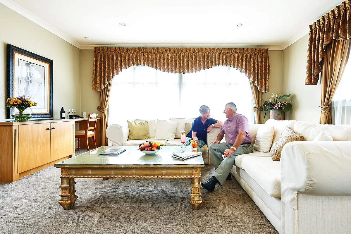 Hotel suite living area featuring white sectional sofa, antique coffee table and couple sharing refreshments.