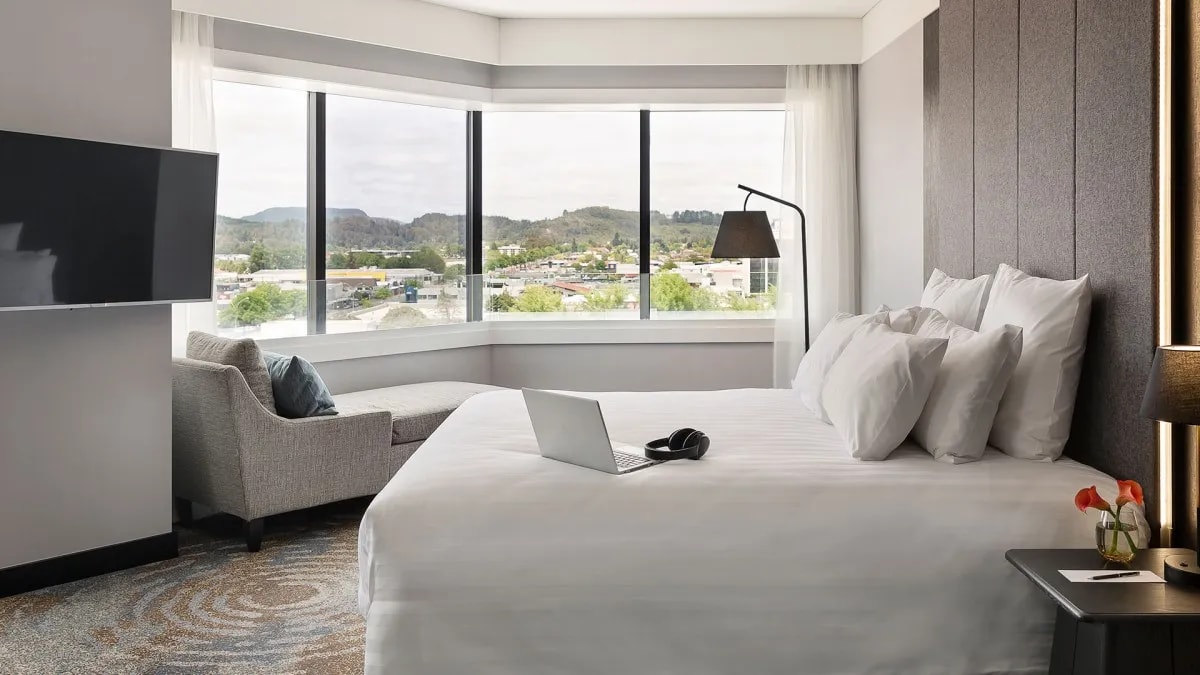Stylish hotel suite with white linens, grey furnishings, wall-mounted TV and scenic views through large windows.