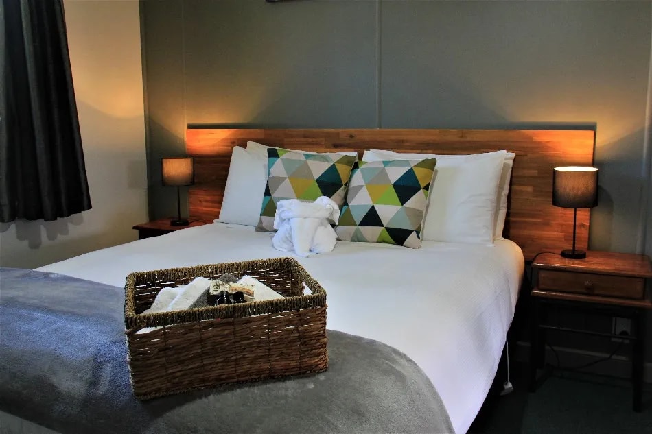 Modern accommodation featuring timber bed frame, colourful pillows, bedside lighting and welcome basket.