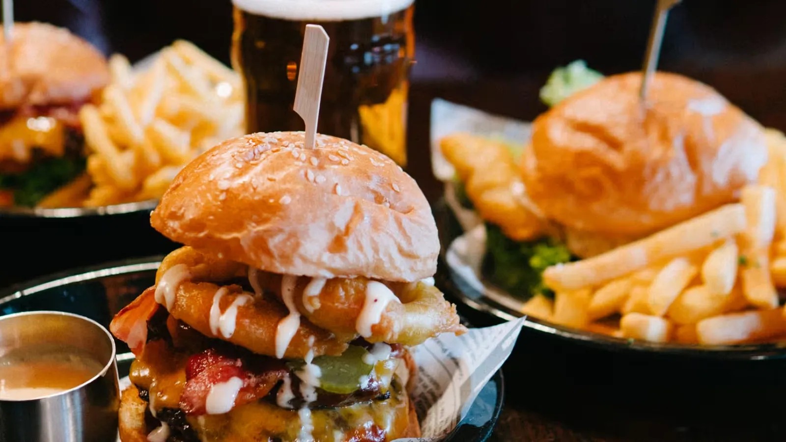 Three burgers in bowls surrounding a glass of beer. The closest burger features onion rings inside, while the other two burgers are accompanied by fries.