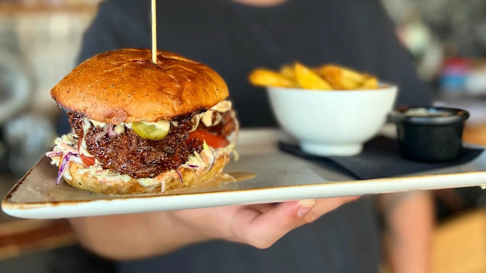 Person holding a serving tray with a loaded beef burger and a bowl of fries and sauce.