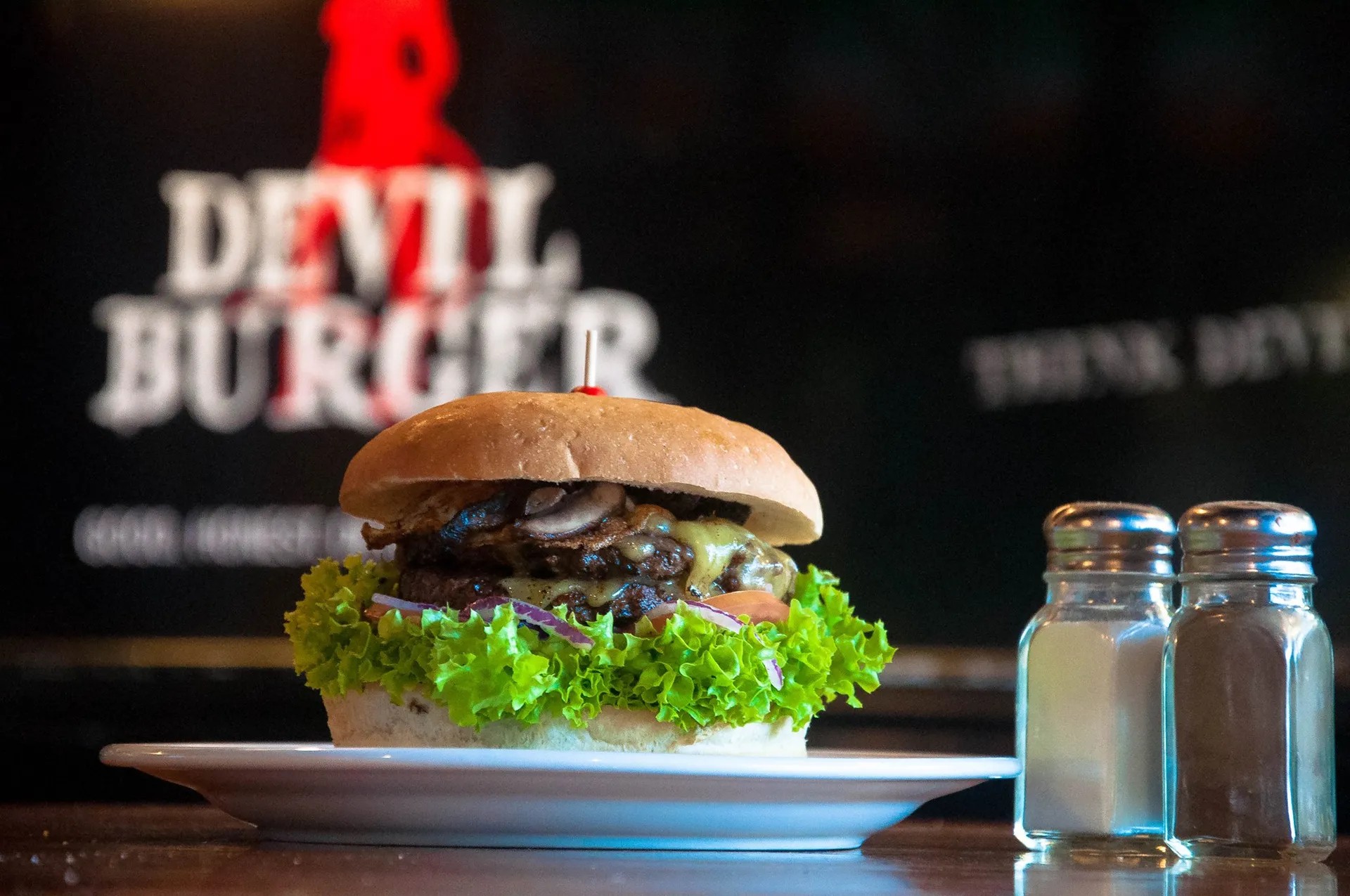 Double beef patty and mushroom burger next to a salt and pepper shaker in front of a black wall with a logo reading "DEVIL BURGER".