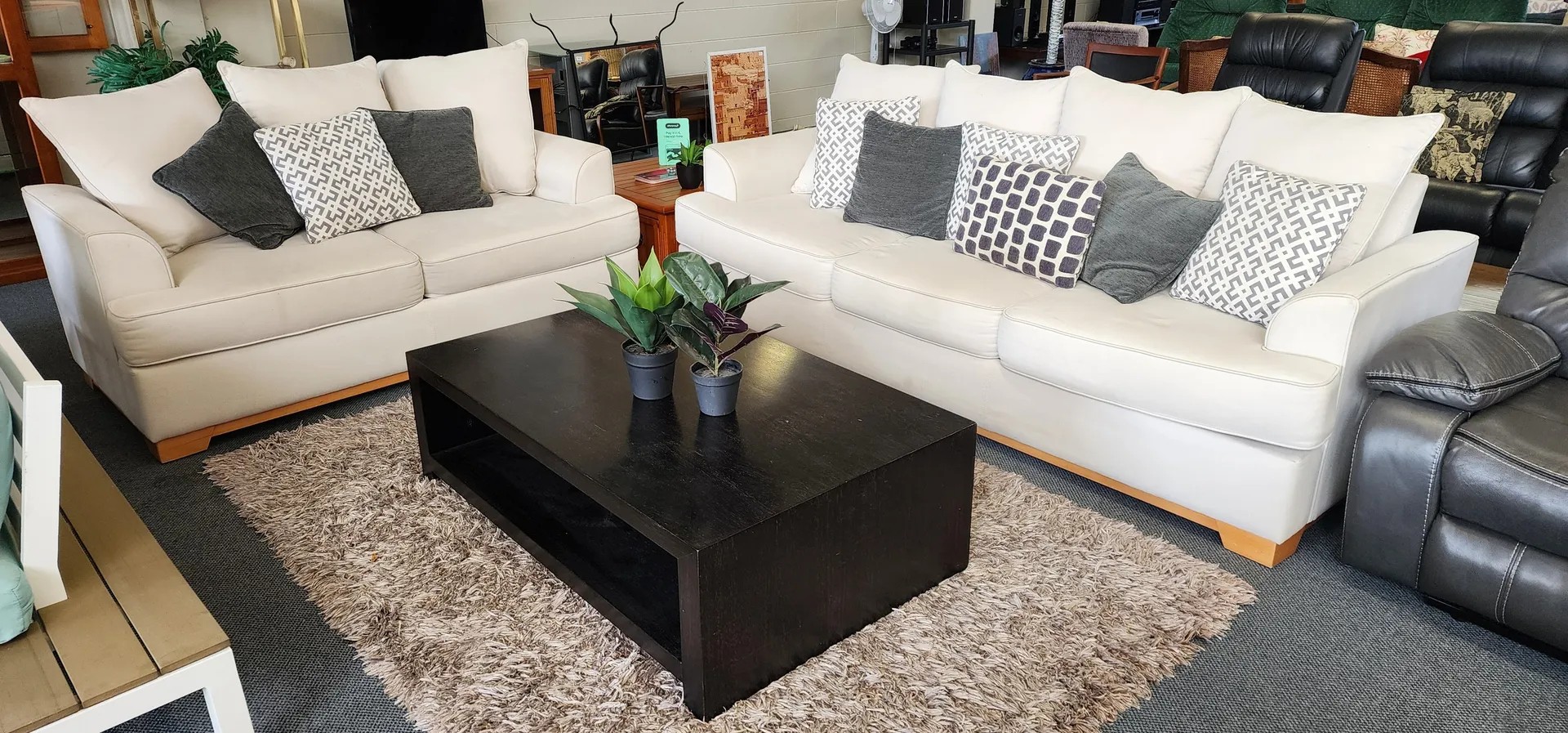 Second-hand white sofa set with decorative throw pillows arranged around dark coffee table in charity furniture shop.