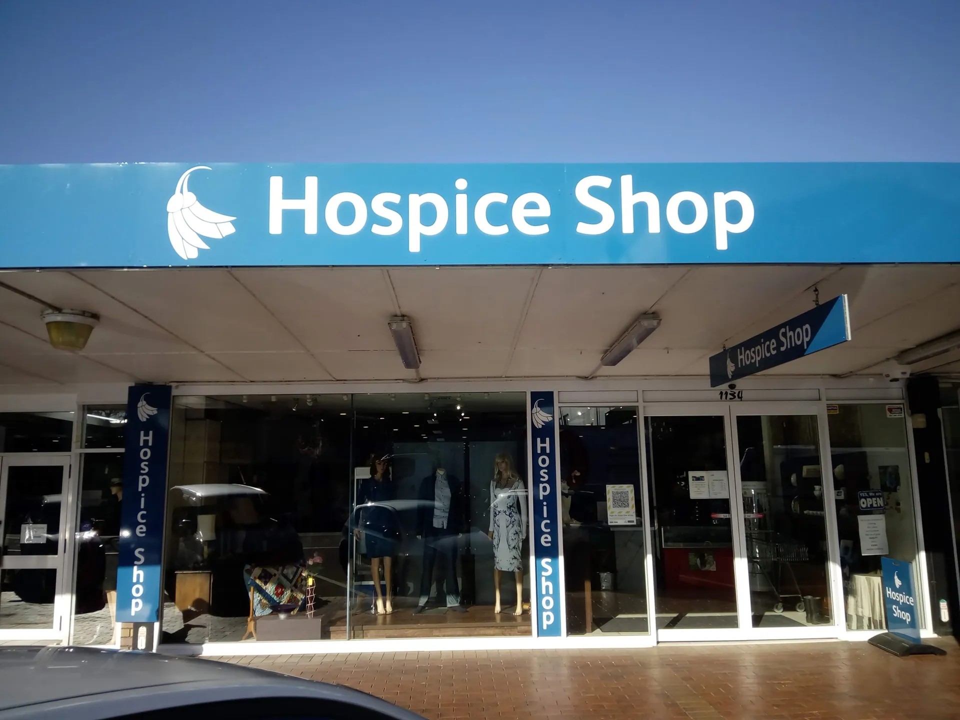 Hospice Shop storefront with blue signage, displaying clothing and goods in glass-fronted windows.