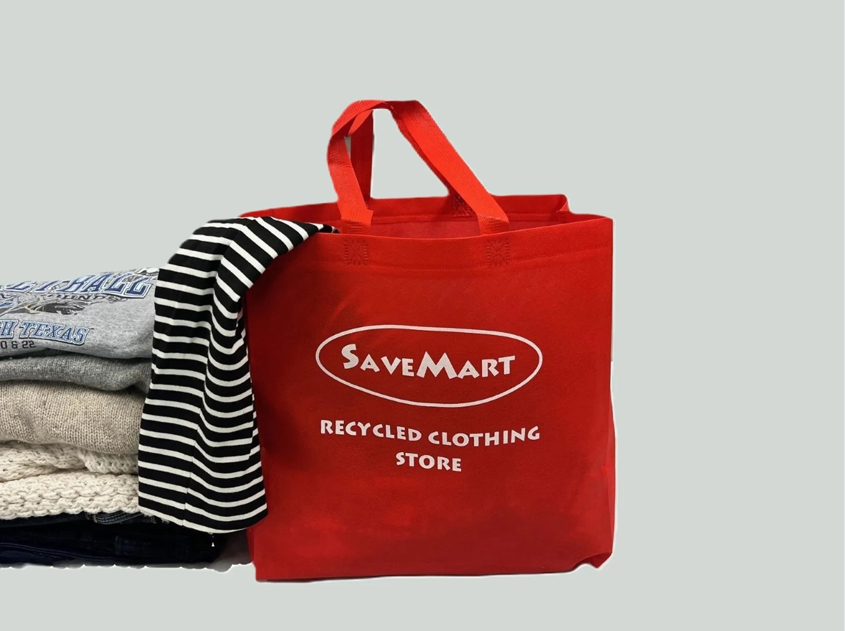 SaveMart reusable shopping bag in red with white branding, containing stack of second-hand clothes and striped fabric.