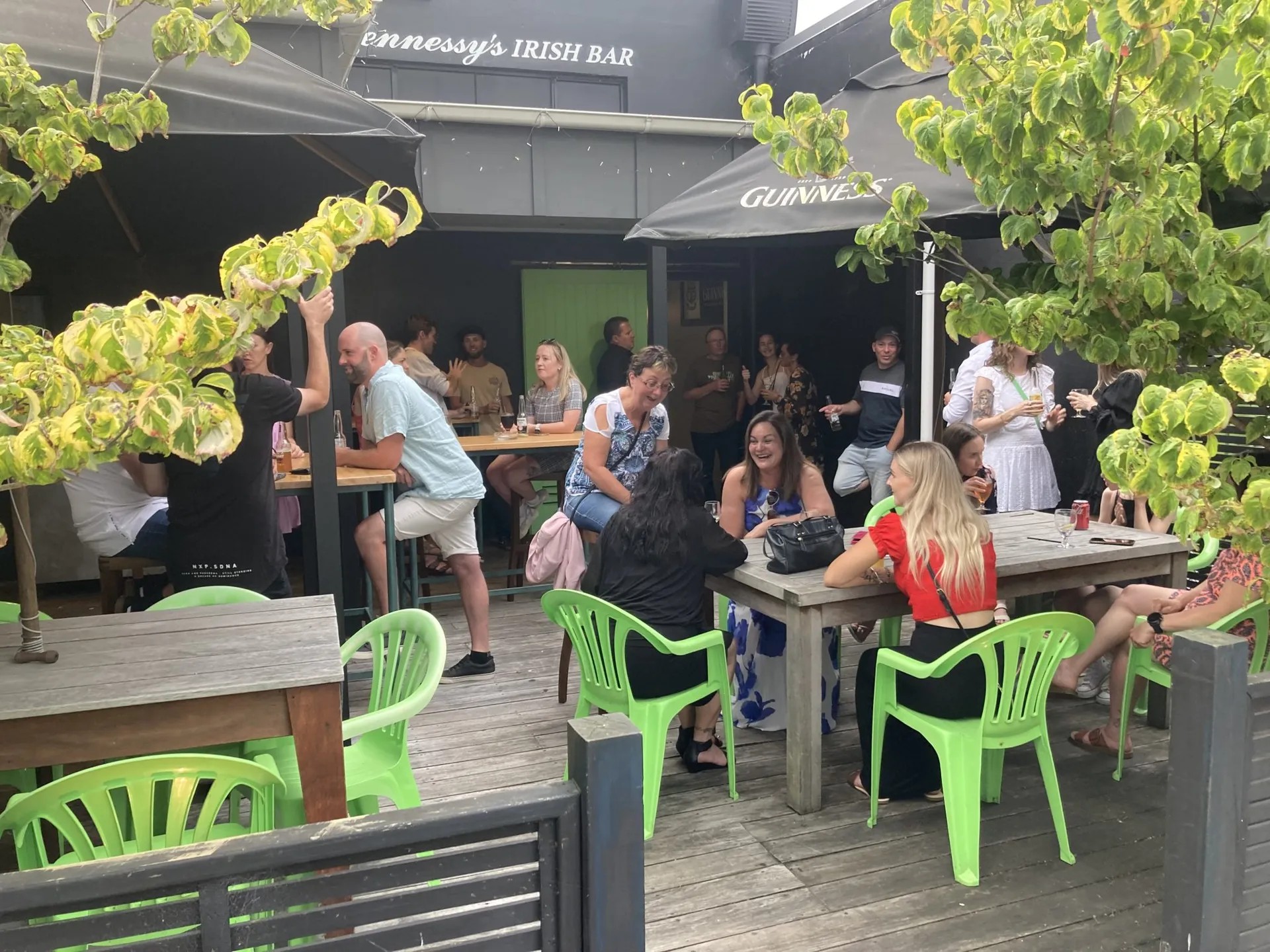 Many customers sitting outside on bright green chairs on the deck of a pub with signage reading "Hennessy's Irish Bar".