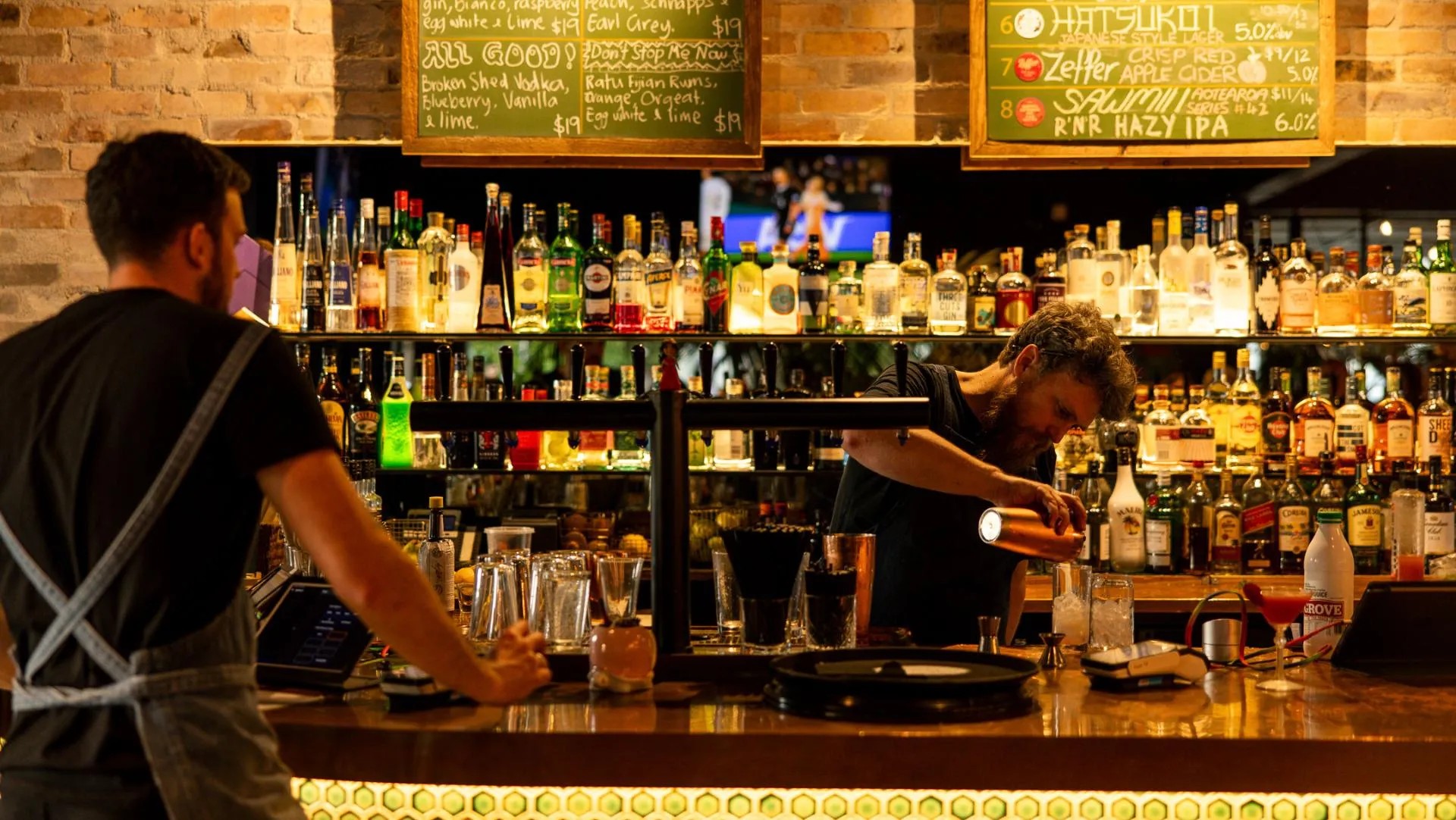 A server stands at the outside a bar counter while a bartender prepares a cocktail on the inside.