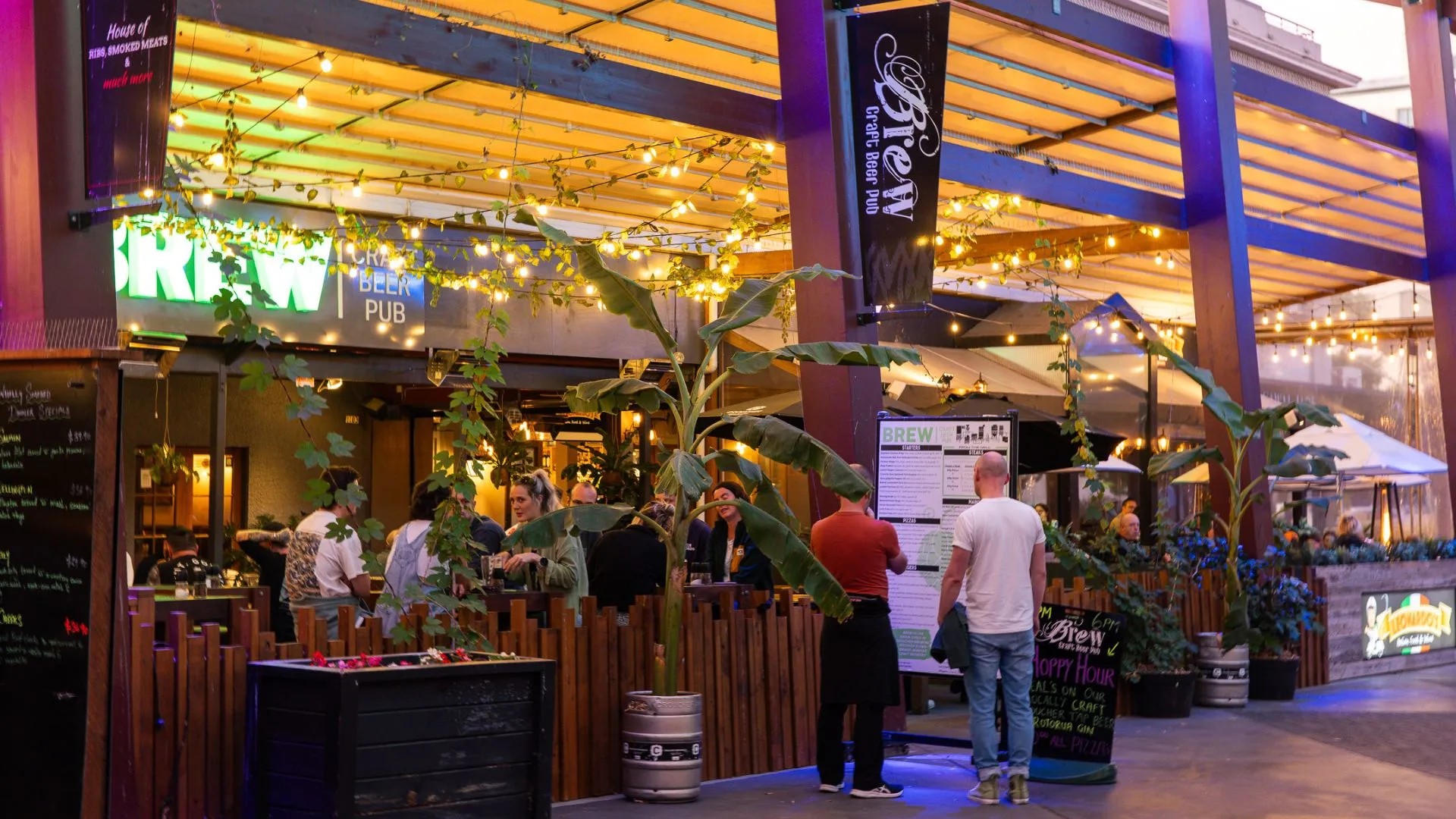 The exterior of a brightly-lit pub with signage reading "Brew Craft Beer Pub". Two people read the menu on the outside of a fence while multiple groups of customers are eating inside.