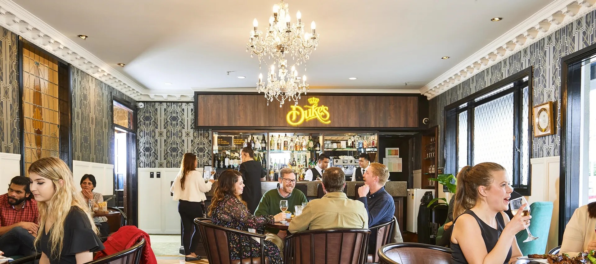 Groups of customers drinking at tables in a high-class pub in front of a bar stand with signage reading "Dukes's".