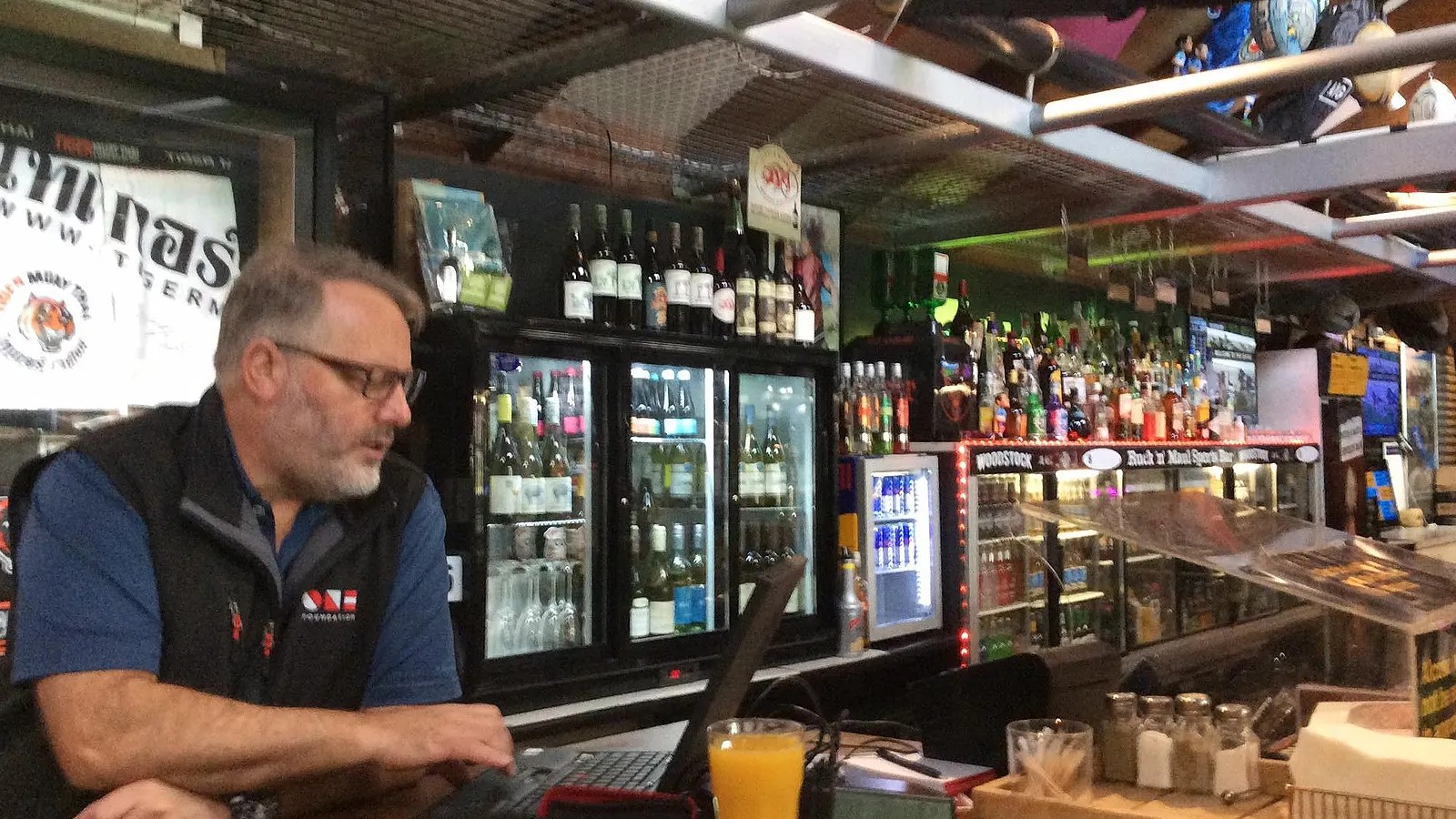 A man uses a laptop on a bar counter in front of fridges of wine and beer in a brightly-lit pub.