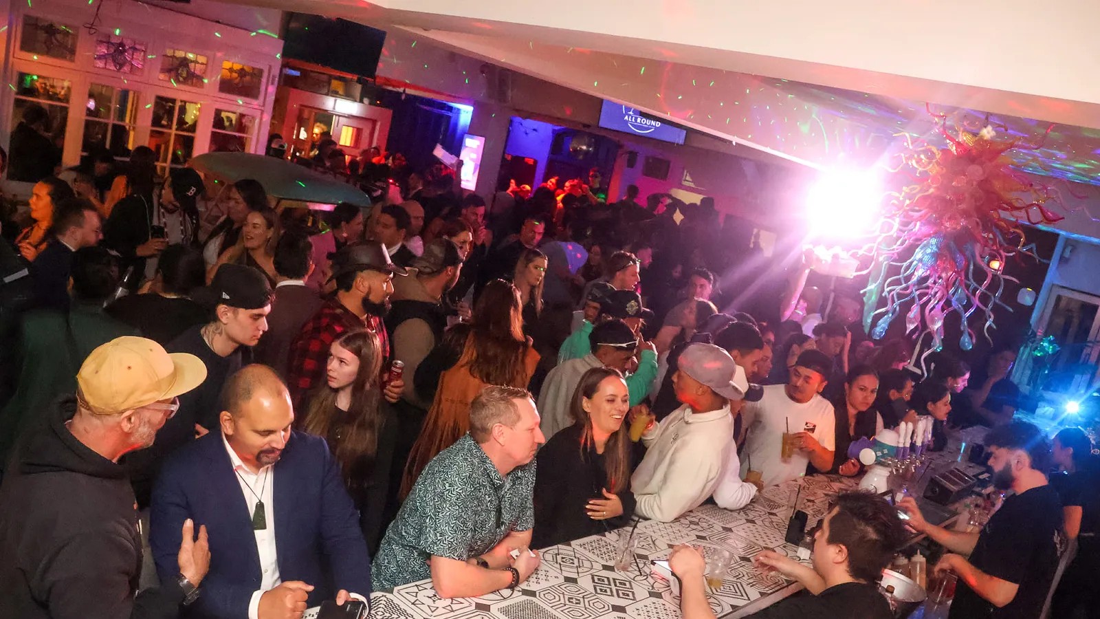 A large crowd of people in a club with colourful lighting, some ordering drinks at the bar stand.