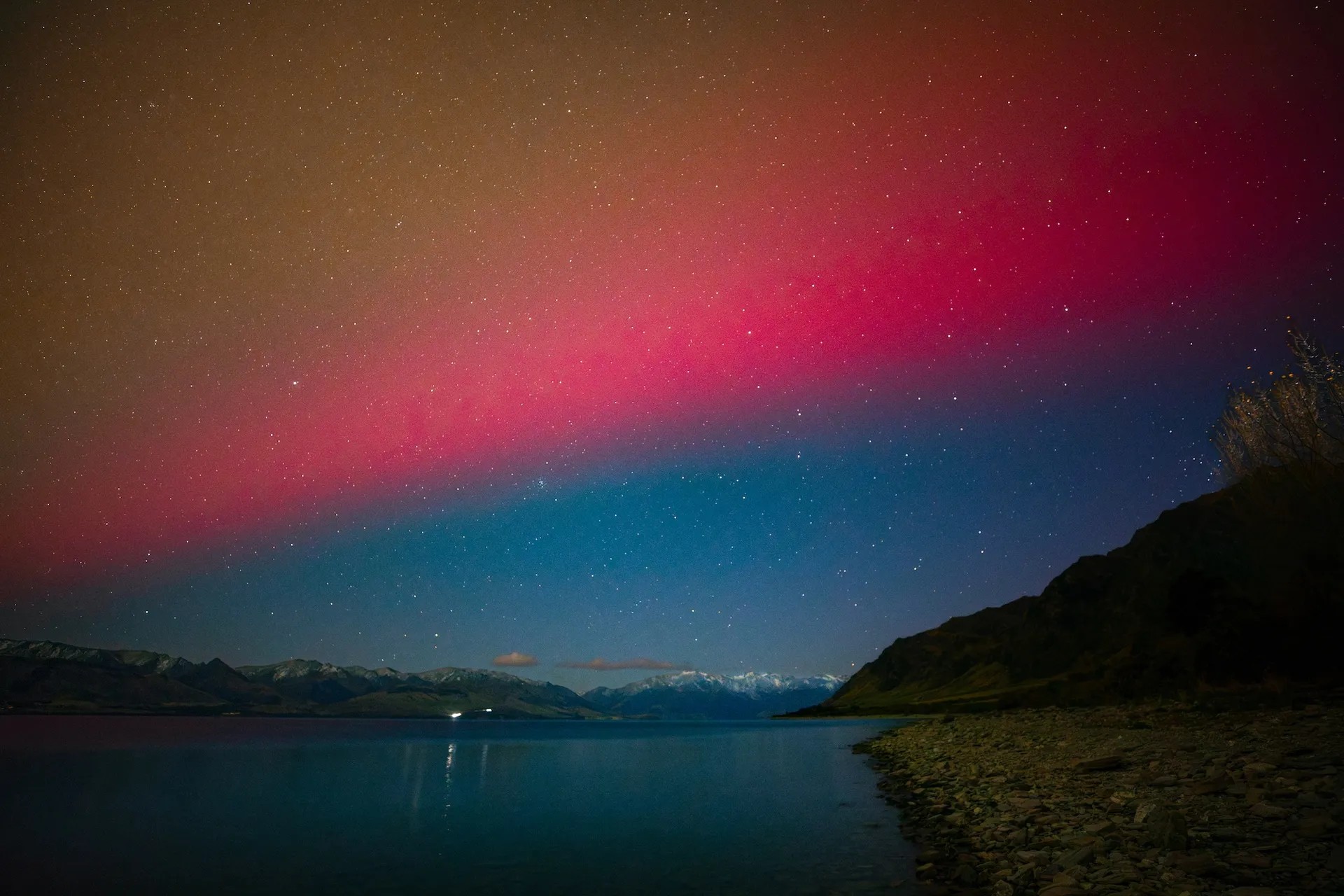 Yellow, pink and blue Southern Lights above water with snowy mountains in the distance.
