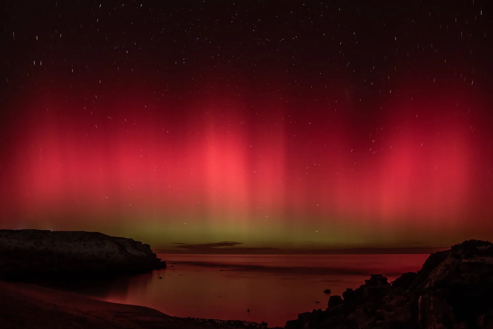 Dark pink, red, and green Southern Lights at rocky beach with stars.