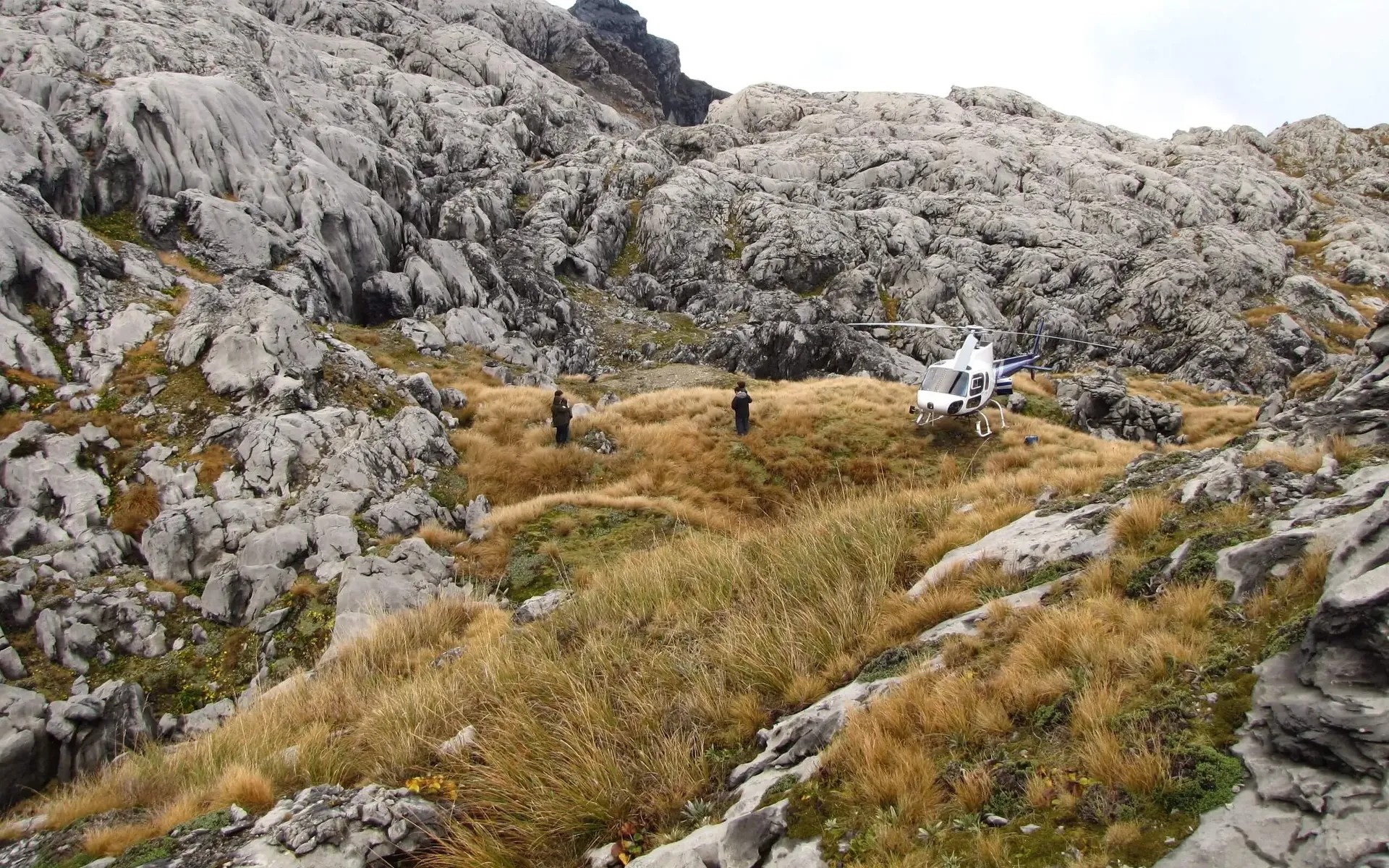 Two people and a helicopter in Mount Owen surrounded by a rocky landscape with tall dry grass.