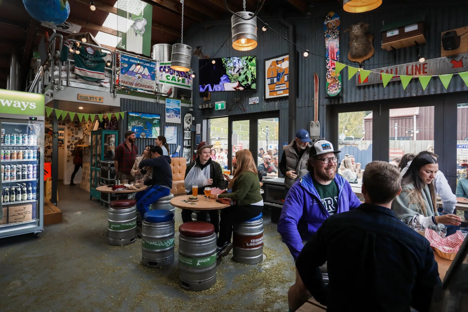 Busy taproom at Altitude Brewing showing beer barrel furniture, hanging keg lights, and customers enjoying drinks.