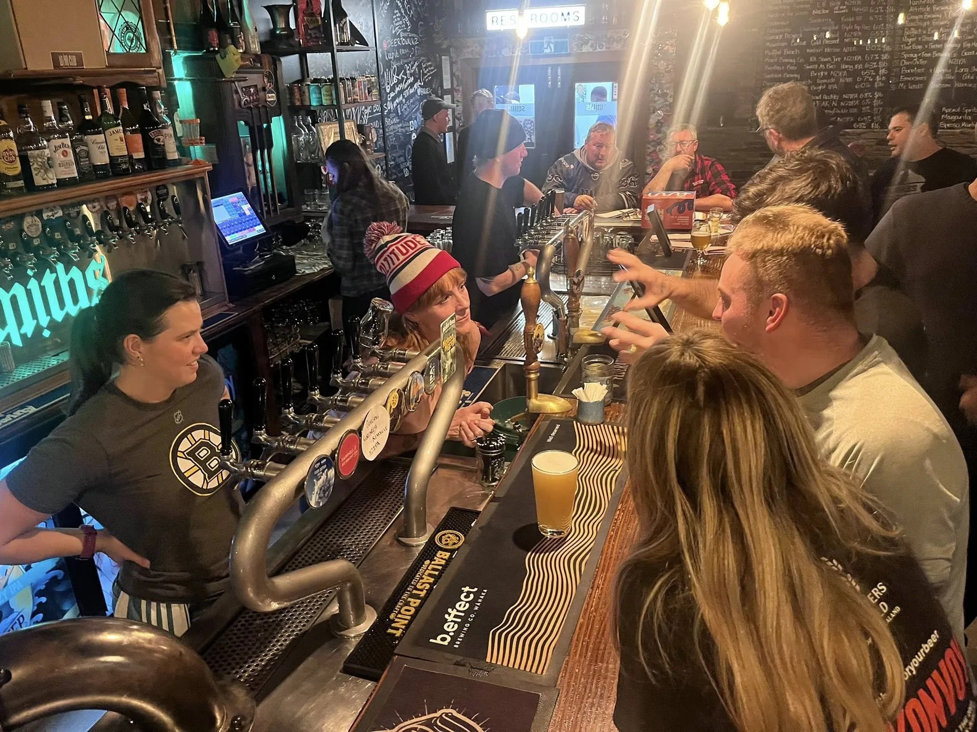 Busy craft beer pub with patrons at the bar, bartender pouring drinks, beer taps, and chalkboard menus on brick walls.