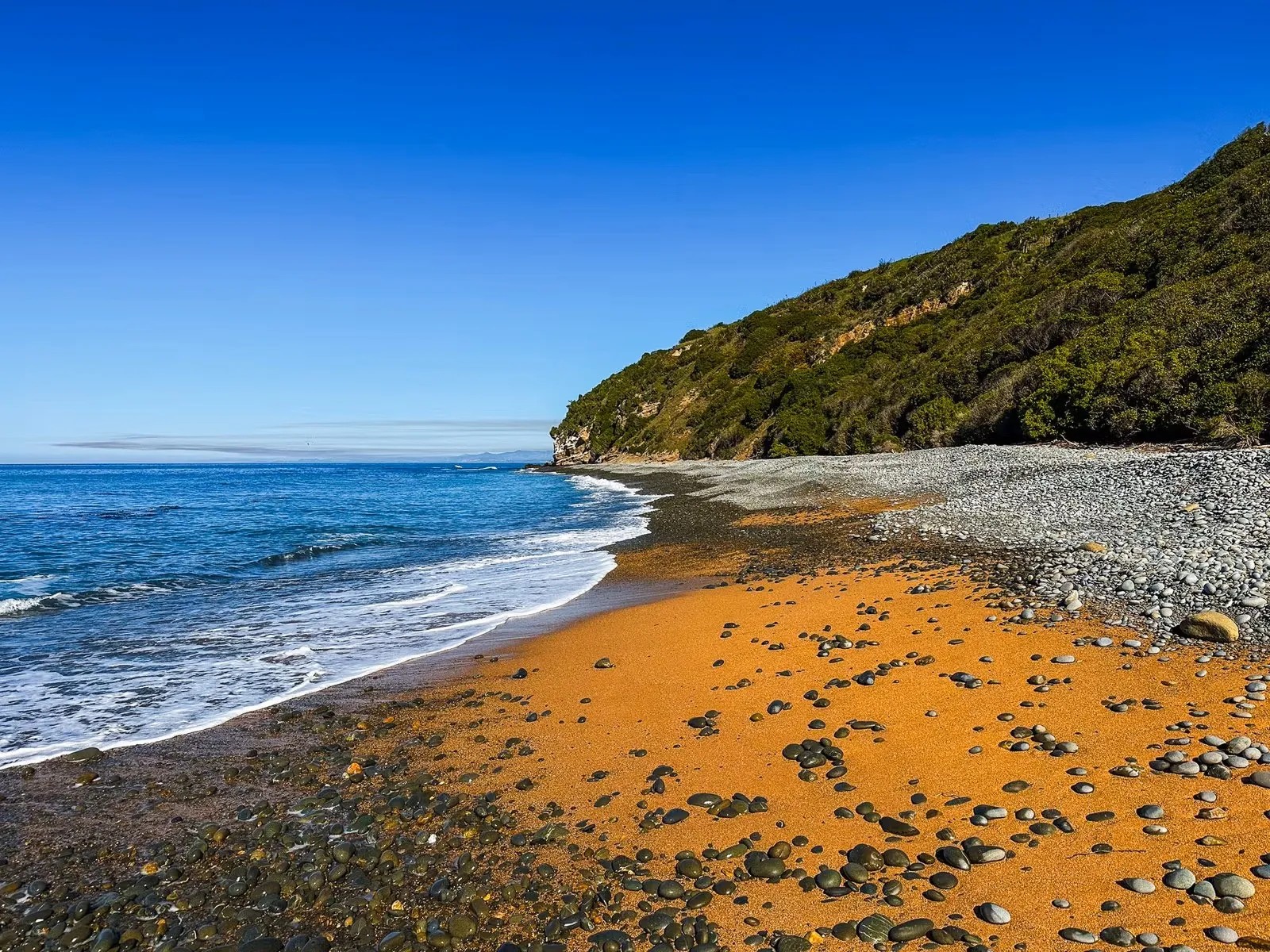 Bushy Beach Oamaru with orange sand, pebbles and clear blue sea water.