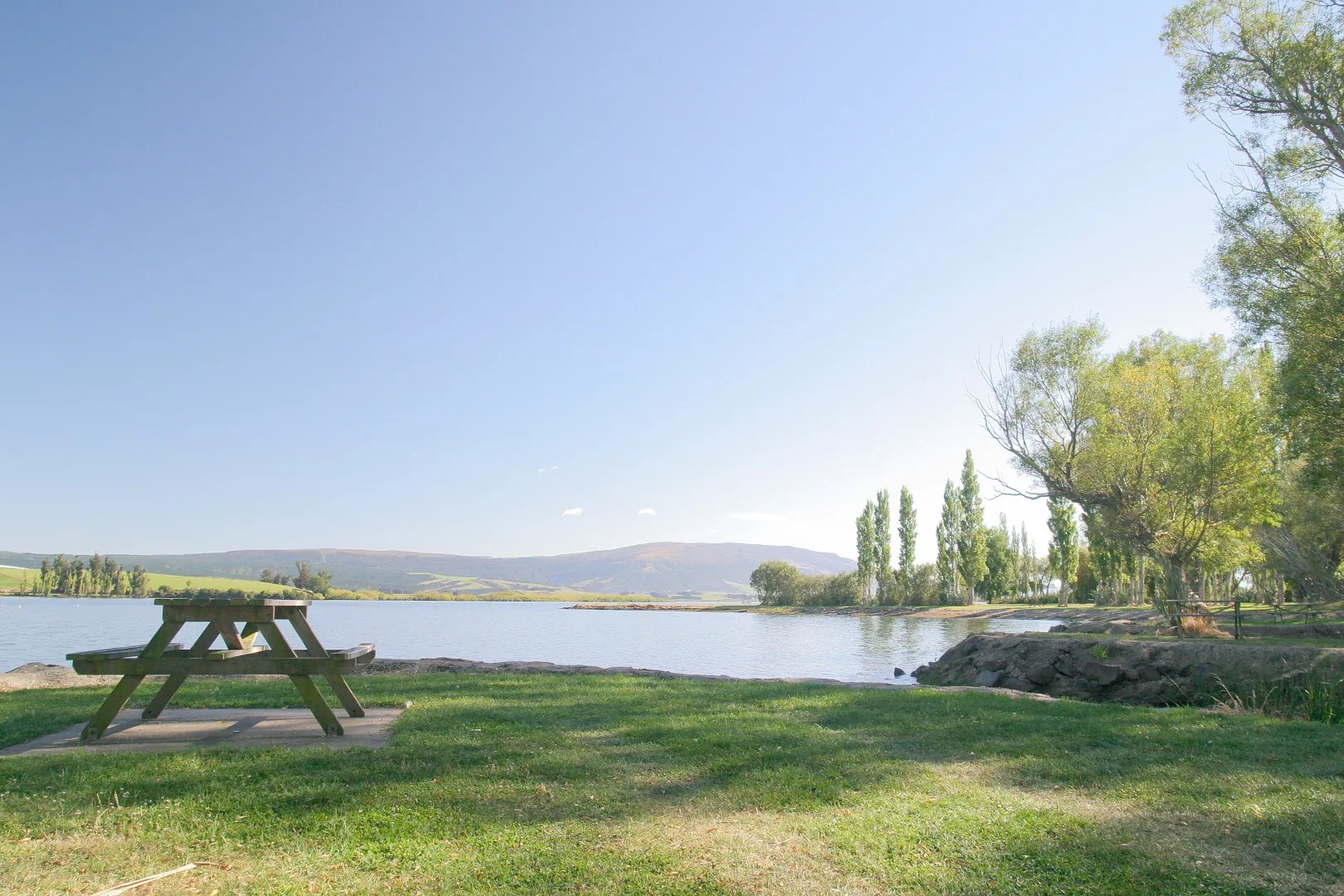 Lake Waihola on a clear day with trees and a picnic table visible close by.