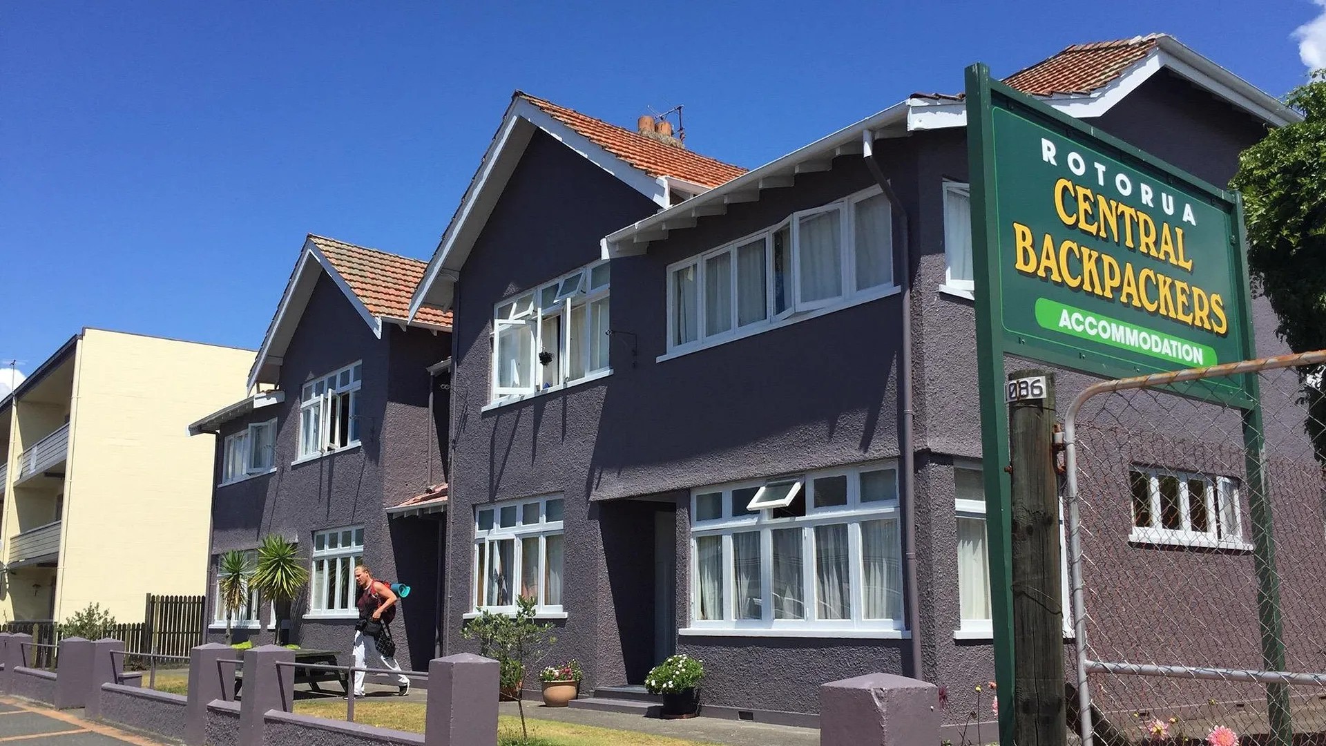 Hostel building with grey walls, white windows, orange roof tiles, and exterior signage reading "Rotorua Central Backpackers Accommodation".
