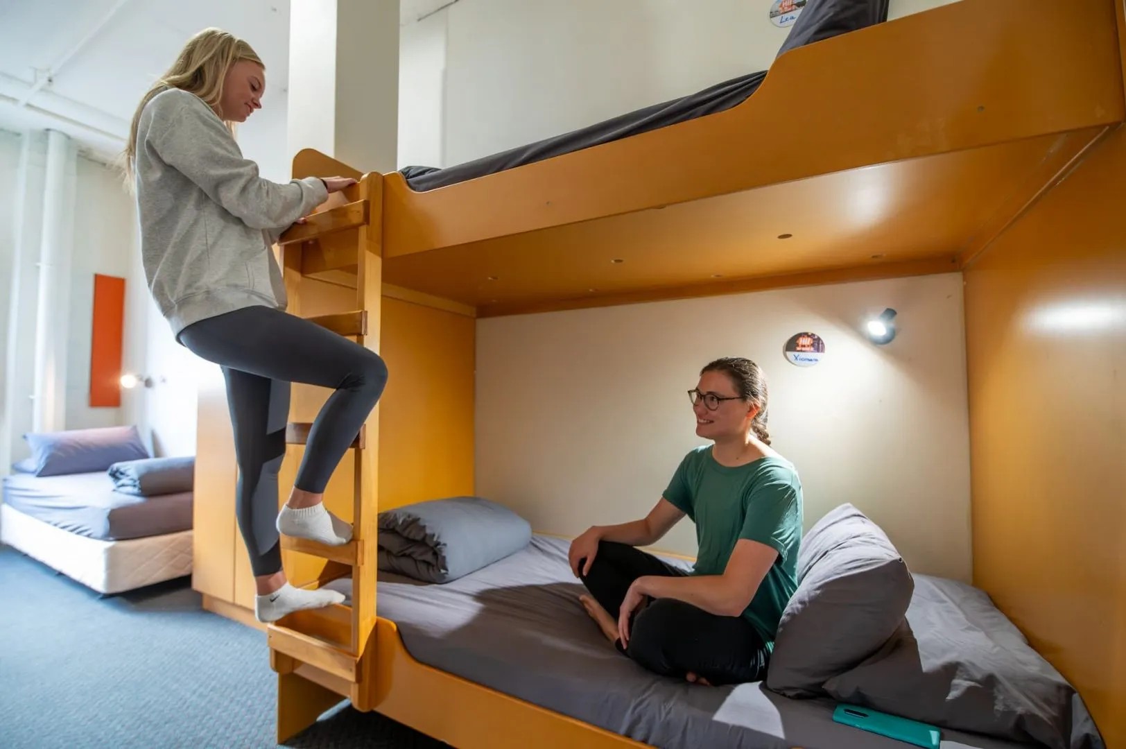 Two backpackers in modern hostel dorm room with wooden bunk beds - one climbing ladder, another relaxing on lower bunk.