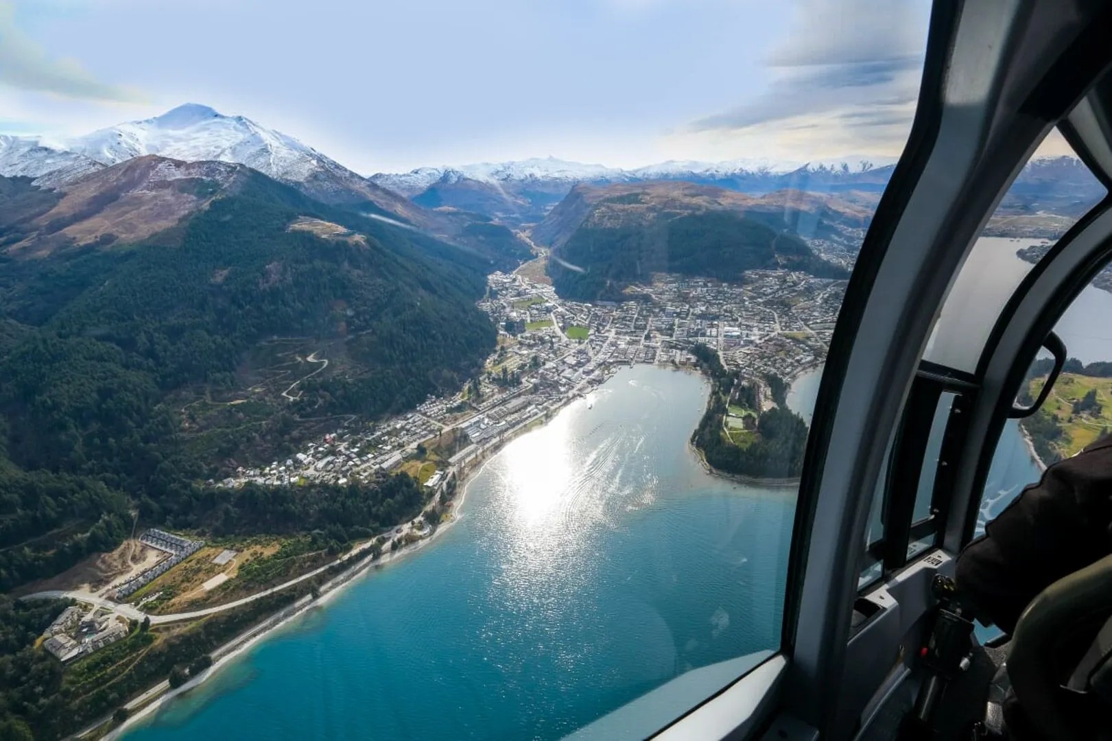 Aerial view of Queenstown from helicopter cockpit showing Lake Wakatipu, town centre and snow-capped mountains beyond.