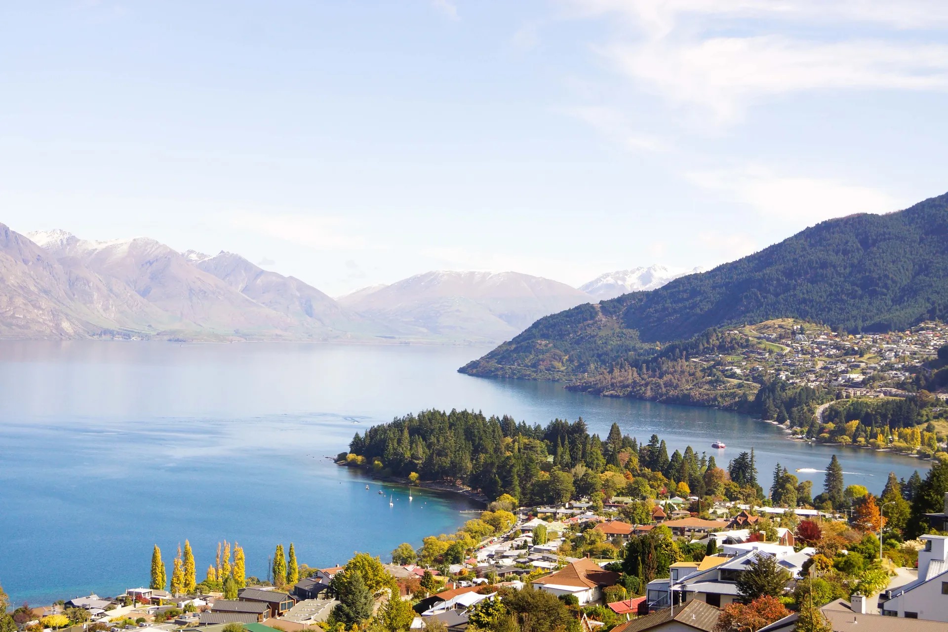 Panoramic view of Lake Wakatipu with Queenstown suburbs, golden autumn foliage and dramatic alpine mountain backdrop.