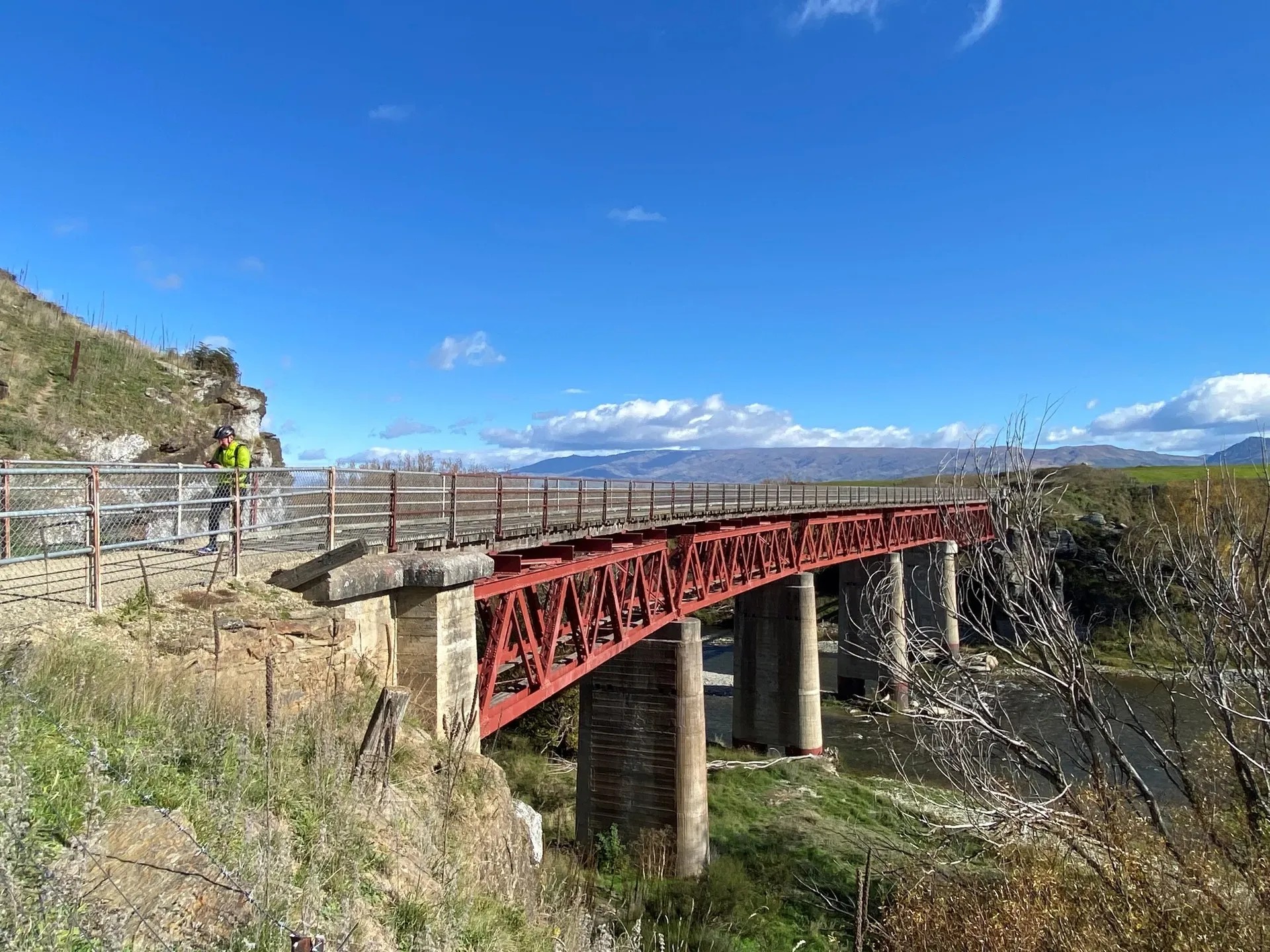 Red metal bridge on the Otago Rail Trail crossing a valley with cyclists and mountain views in Central Otago.