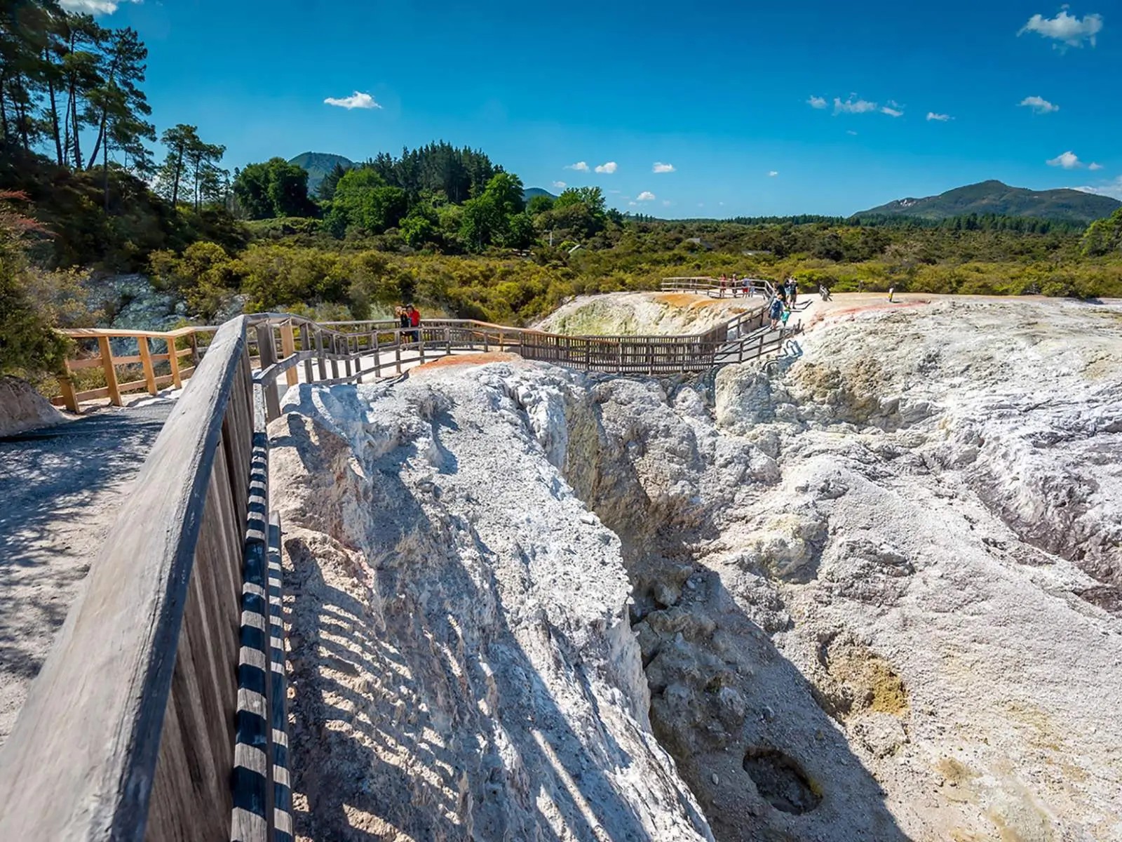 Wooden boardwalks over white geothermal terrain with steam vents at Rotorua, surrounded by native bush and mountains.