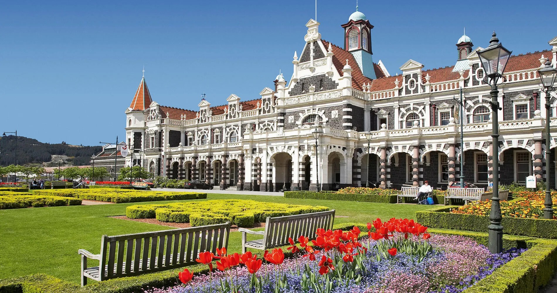 A railway station designed in the Flemish Renaissance architectural style. In front of the station is a well-manicured garden with small hedges and flowers.