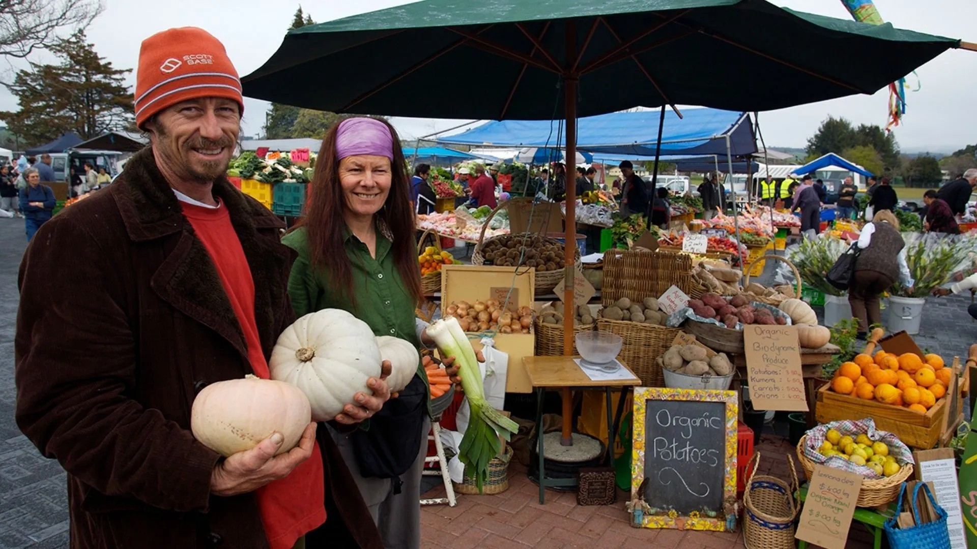 Two people holding vegetable and smiling in front of a produce stand at a farmer's market.
