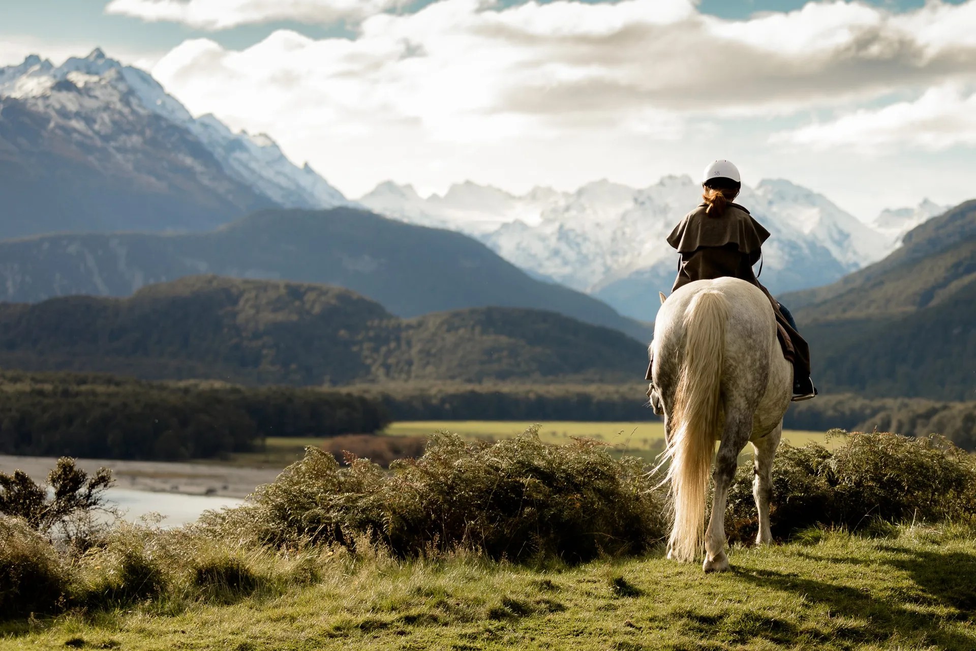 Back view of a person riding a white horse with snowy mountains in the distance.