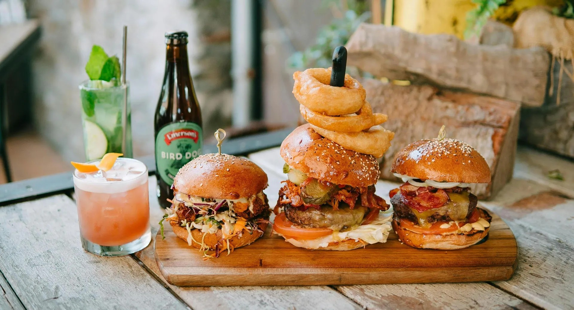 Three different burgers on a wooden board with three drinks next to them.