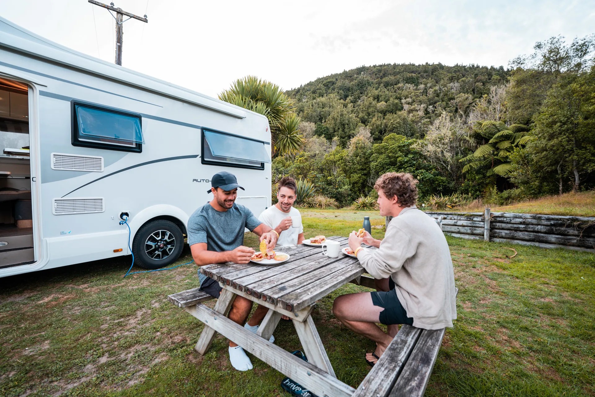 Three people sitting on a picnic bench and eating food in front of a campervan.