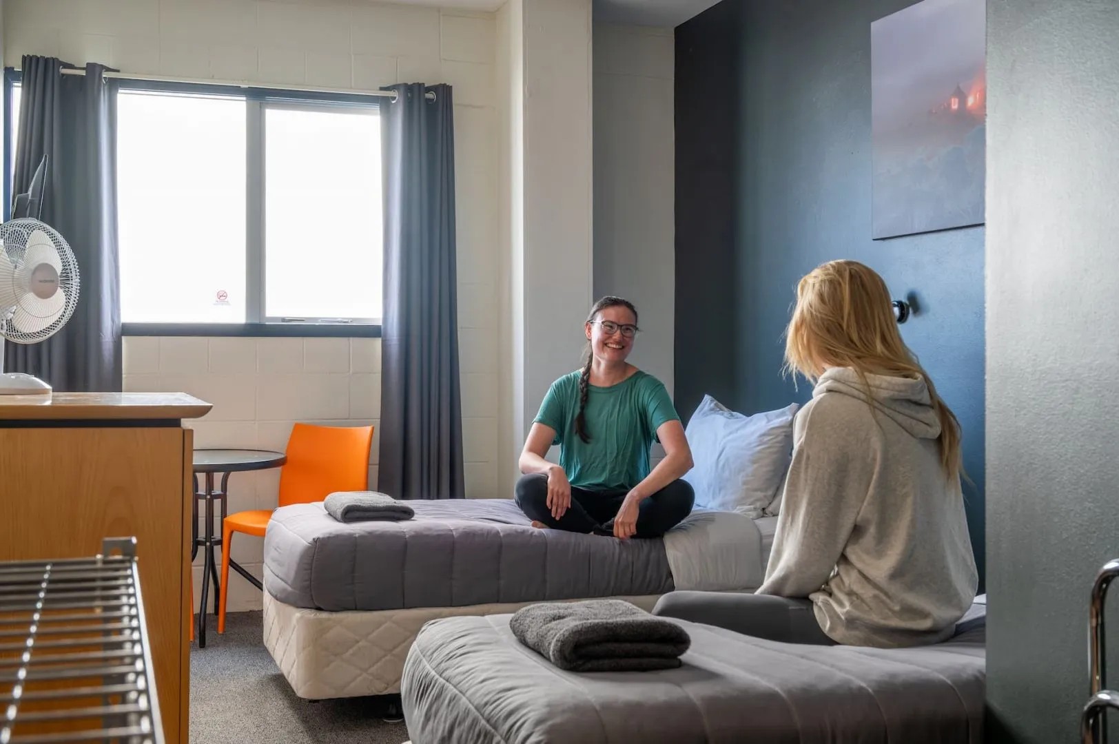 Two people sit on single beds in a small backpacker ensuite room.