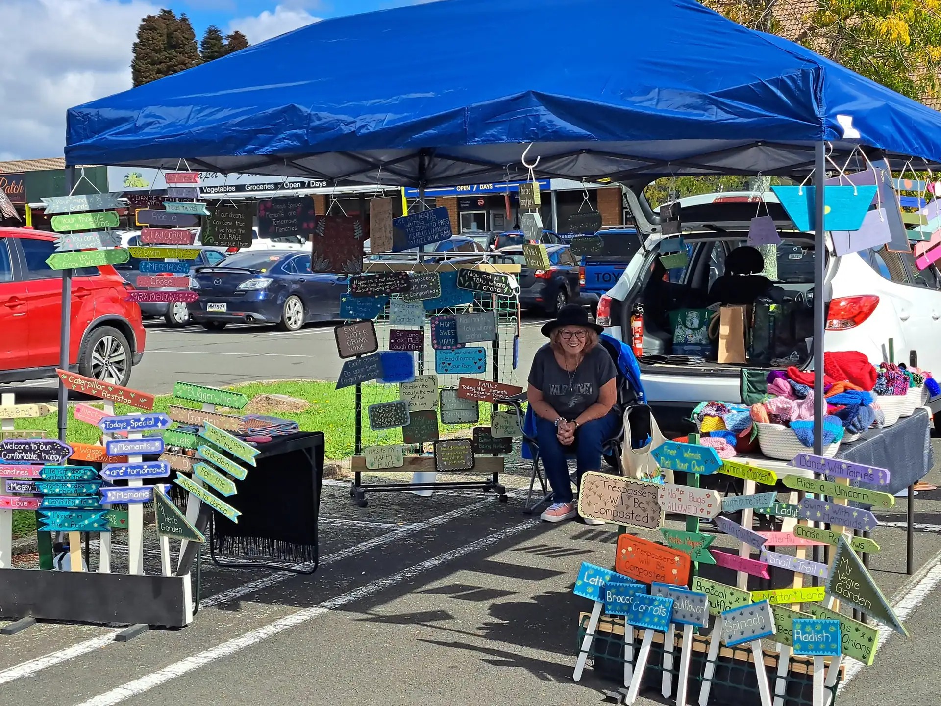 Person sitting under a market tent stall selling wooden signs.