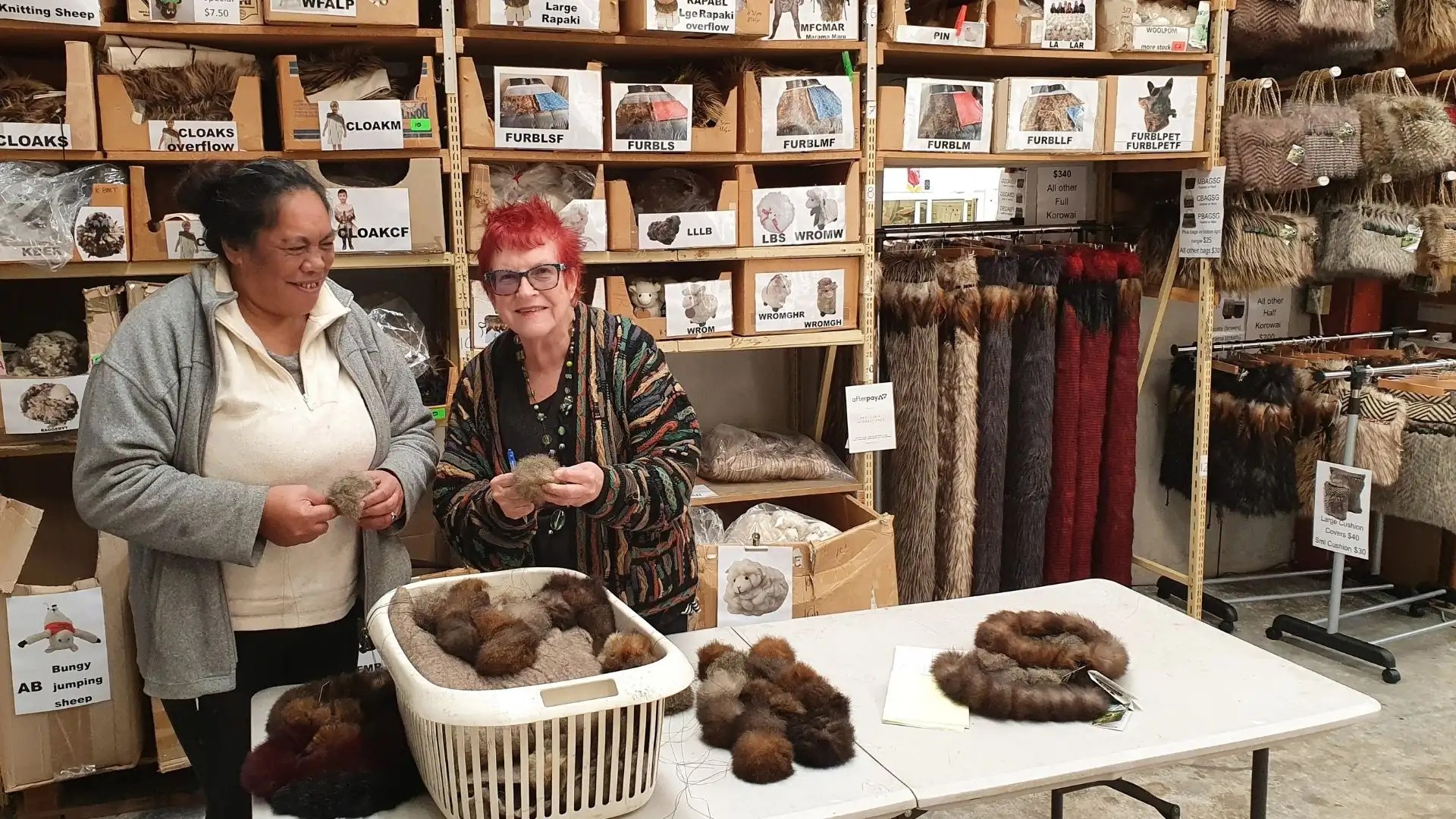 Two employees at a souvenir factory hold possum fur cuffs. Behind them is an array of NZ souvenir products.