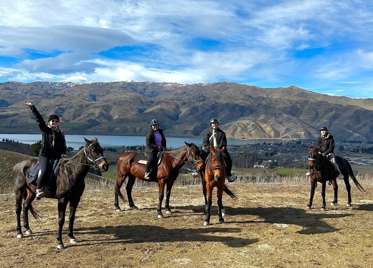 Four people riding horses on a hill, with views of mountains, a lake, and a town in the distance.