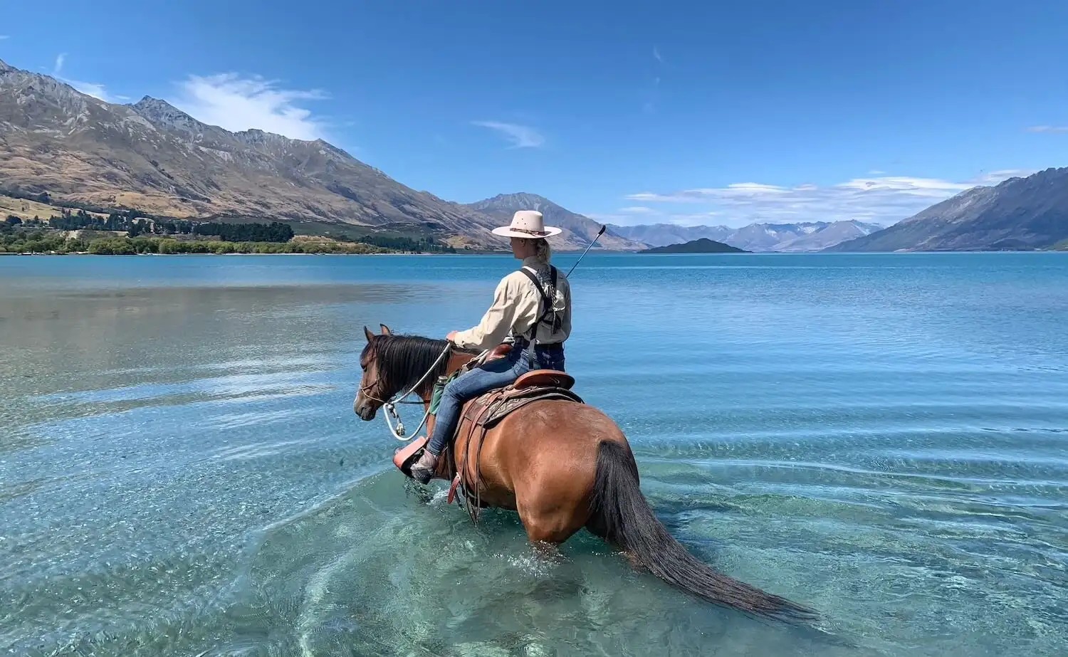 Woman on horse in clear blue lake water looking out at mountains.