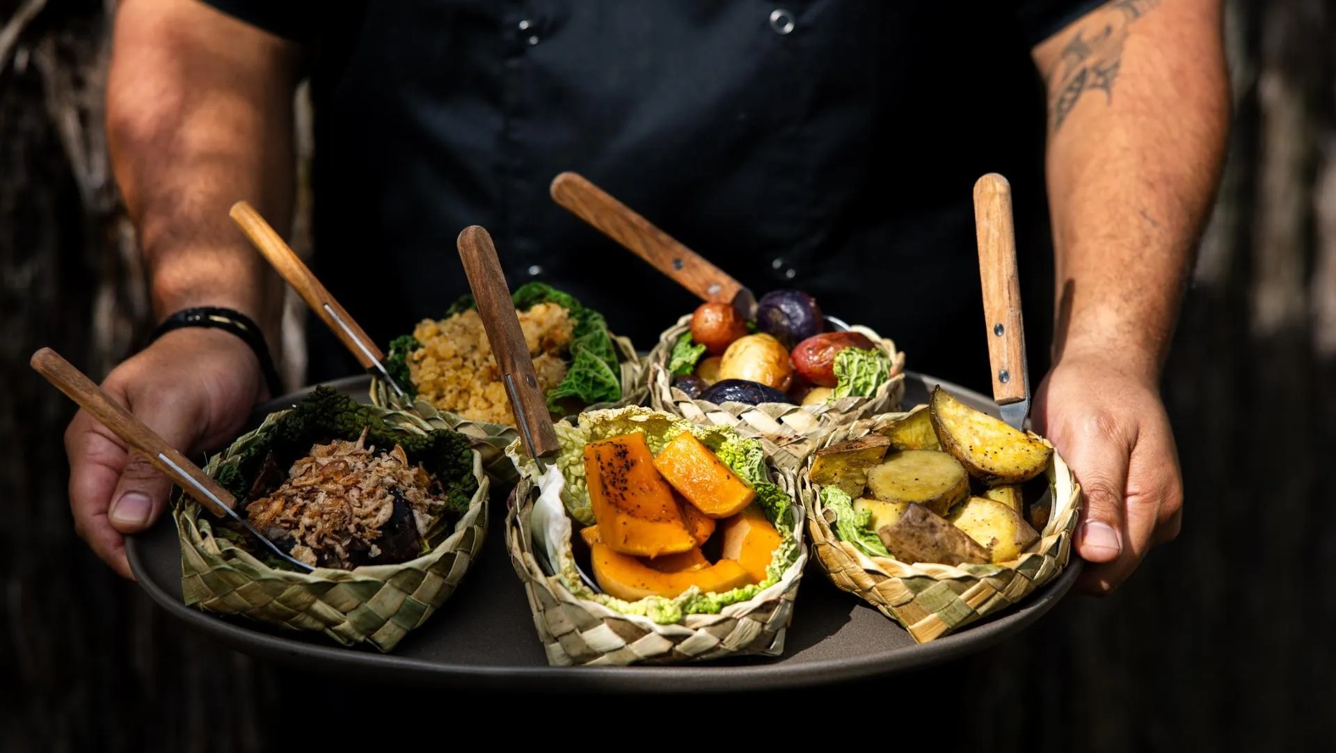 Person holding plate with flax-weaved bowls featuring hangi-inspired fusion food.