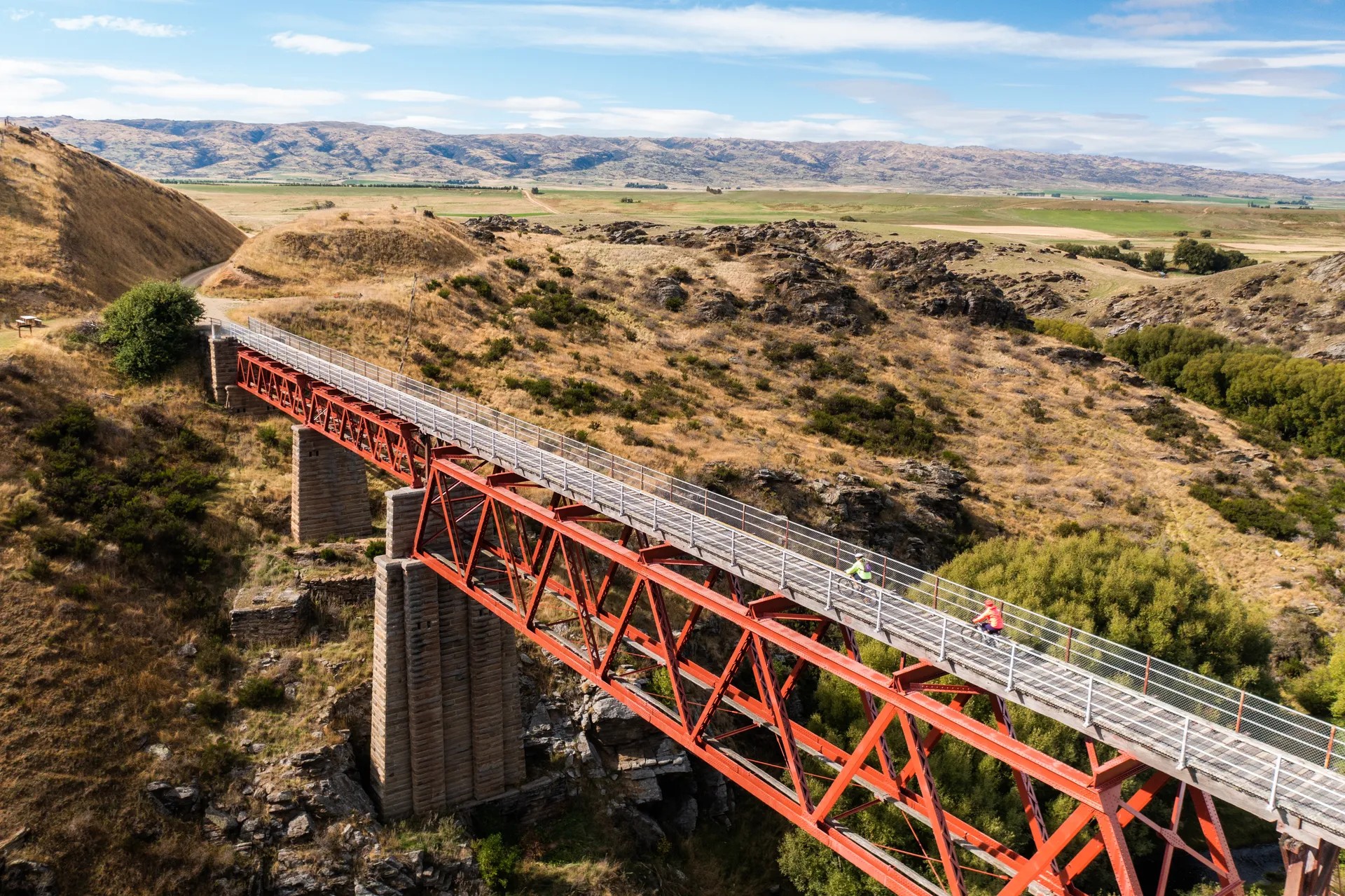 Two cyclists riding across a red railway bridge on grassy, rocky, land.