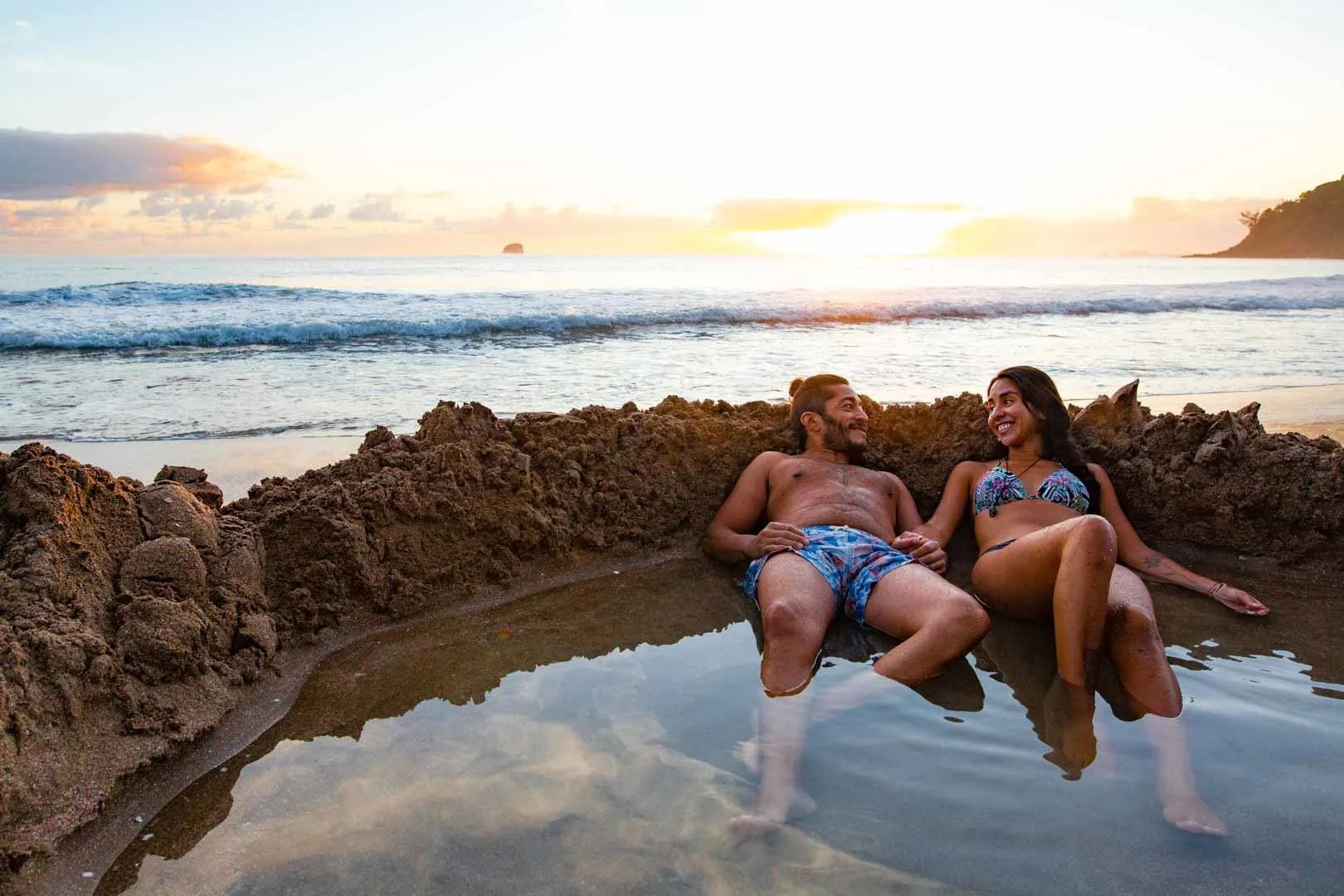 A couple smiling at each other in a large sand pool filled with hot water on a beach.