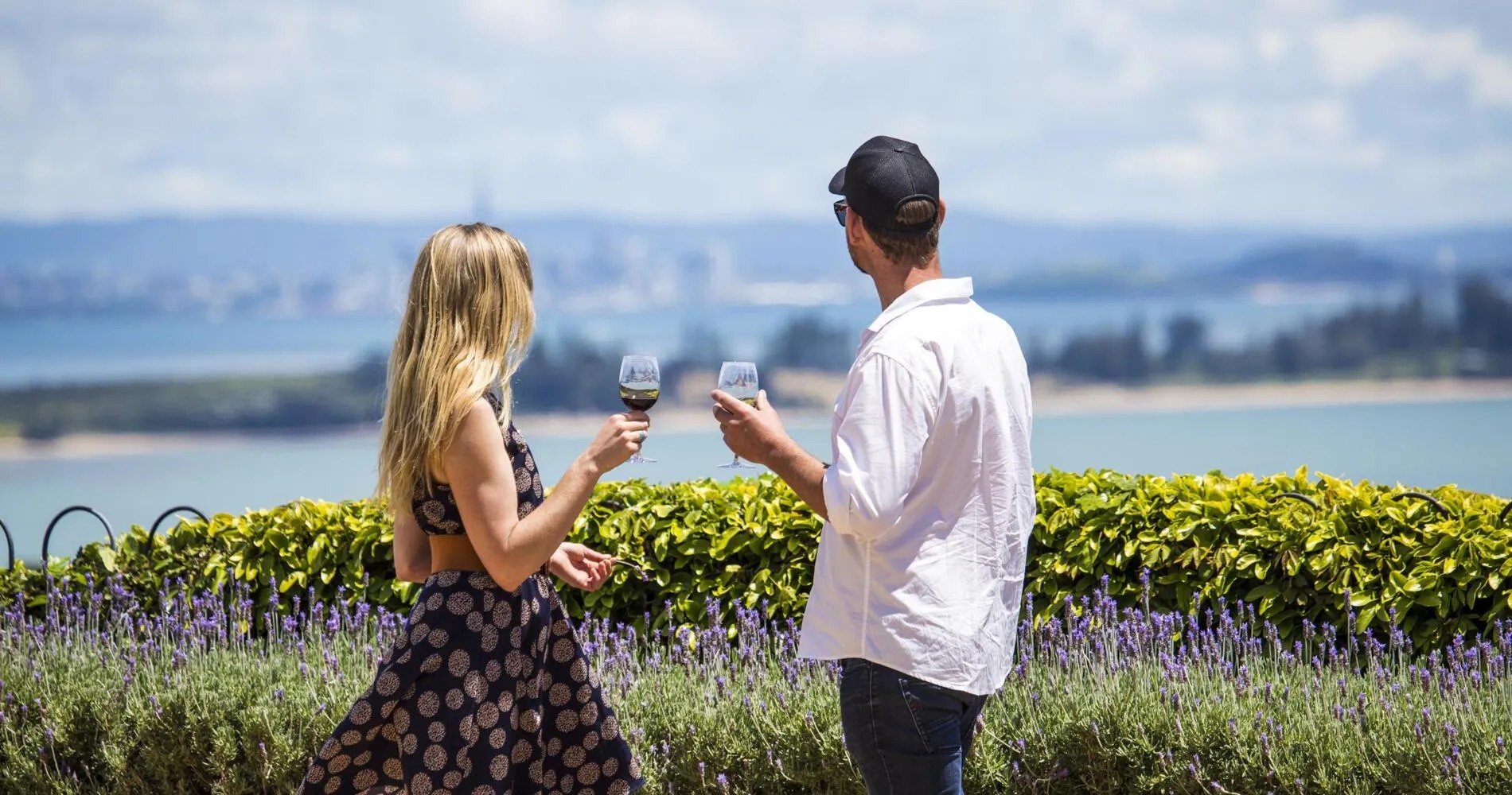 Two people holding white wine glasses as they overlook the sea and distant city.