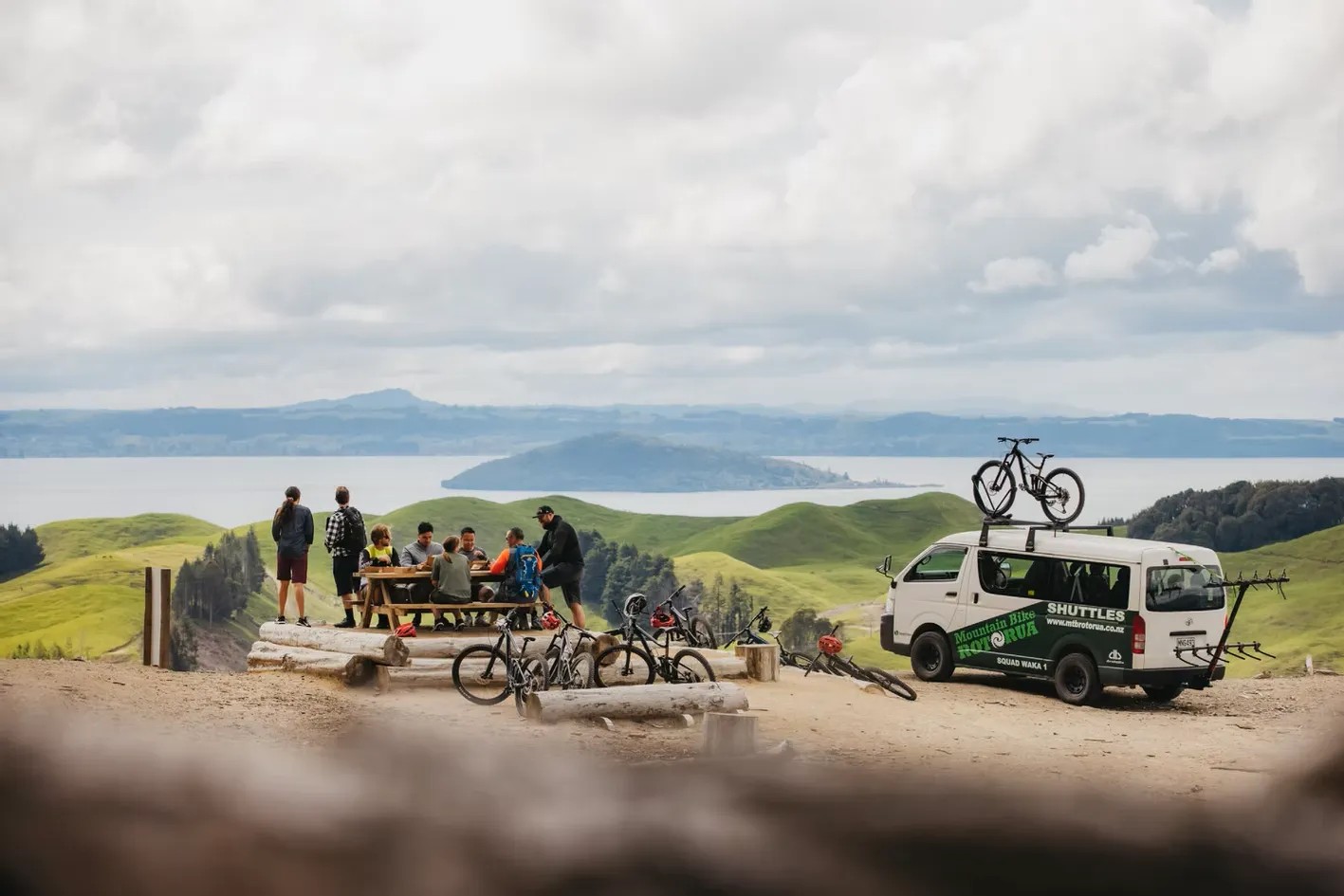 A group of people sitting at a picnic table with a Mountain Bike Rotorua shuttle nearby and Lake Rotorua in the distance.
