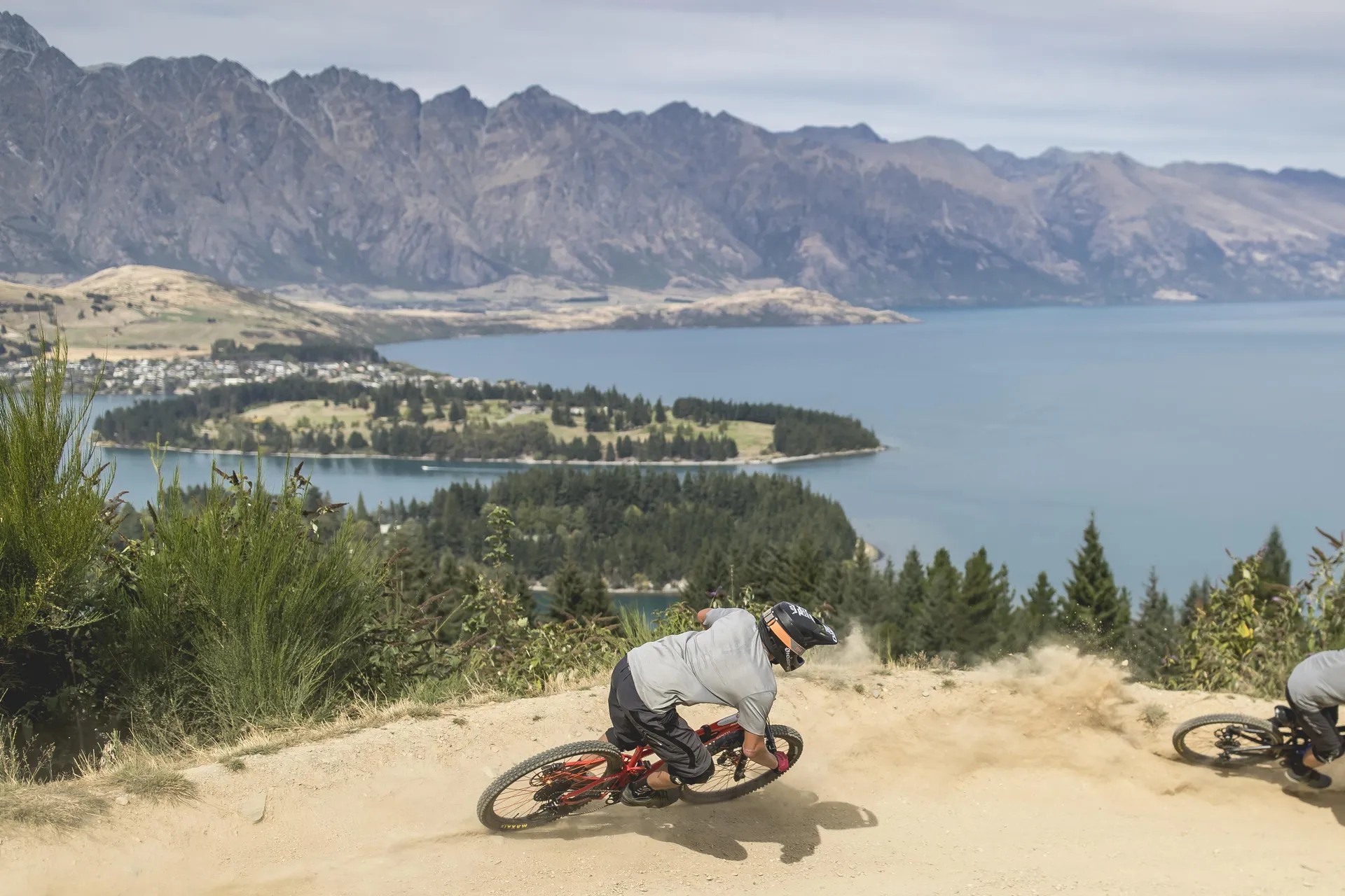 Two cyclists riding along a curve on a path with views of Lake Wakatipu and Queenstown Gardens in the distance.