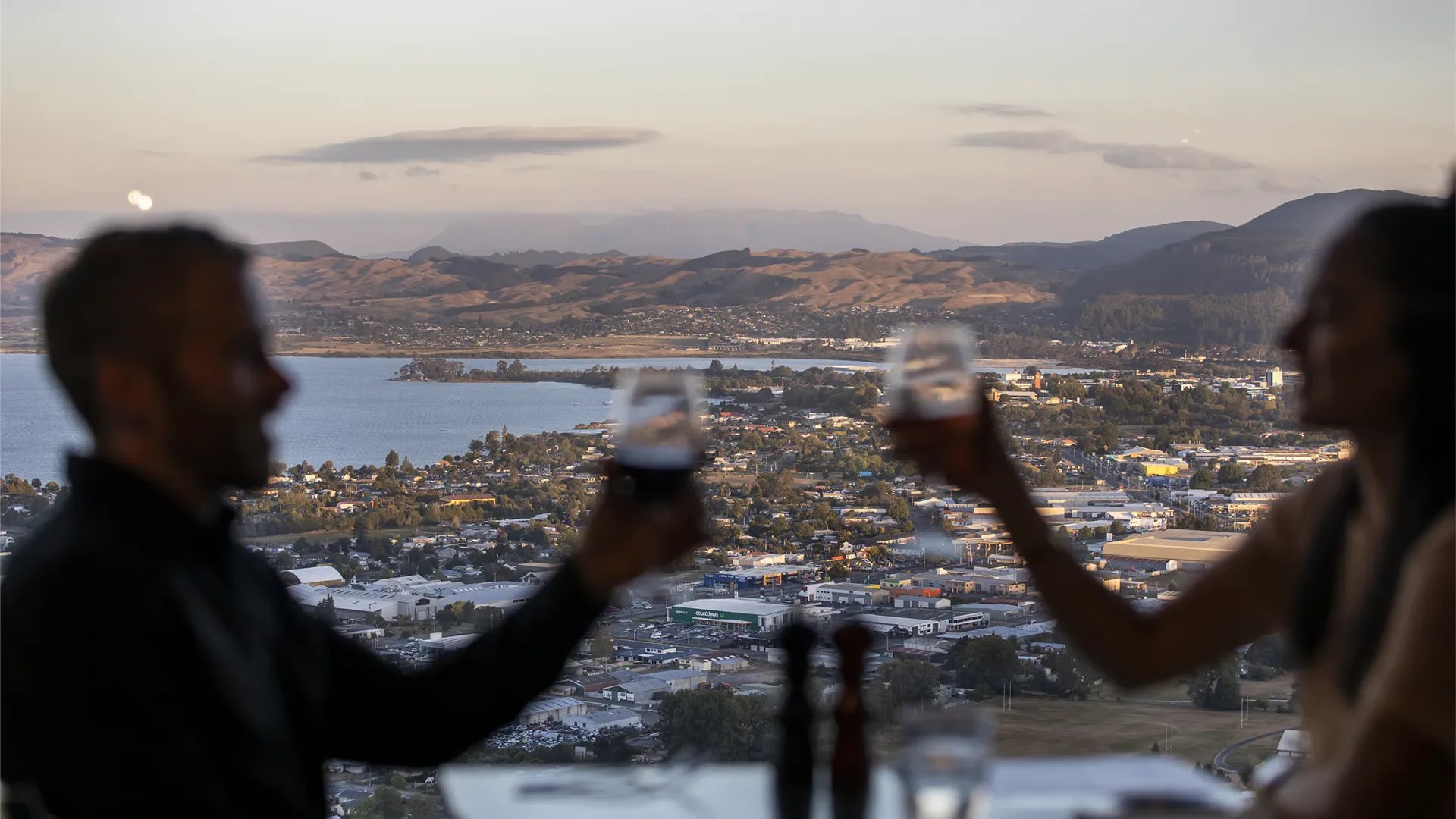 Couple making a toast by the window of a restaurant with views of Lake Rotorua and the city.